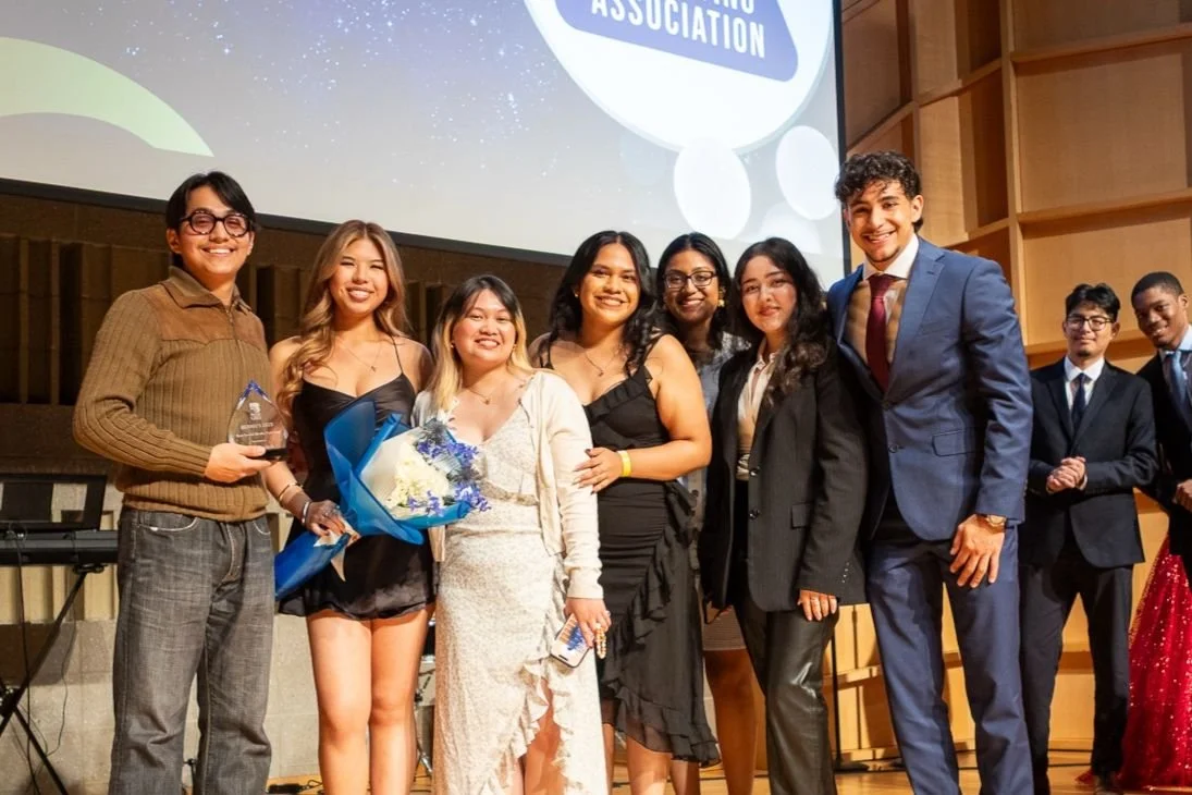 Group of young adults dressed in formal attire standing on stage at an award ceremony, with some holding awards and flowers, smiling for the photo.