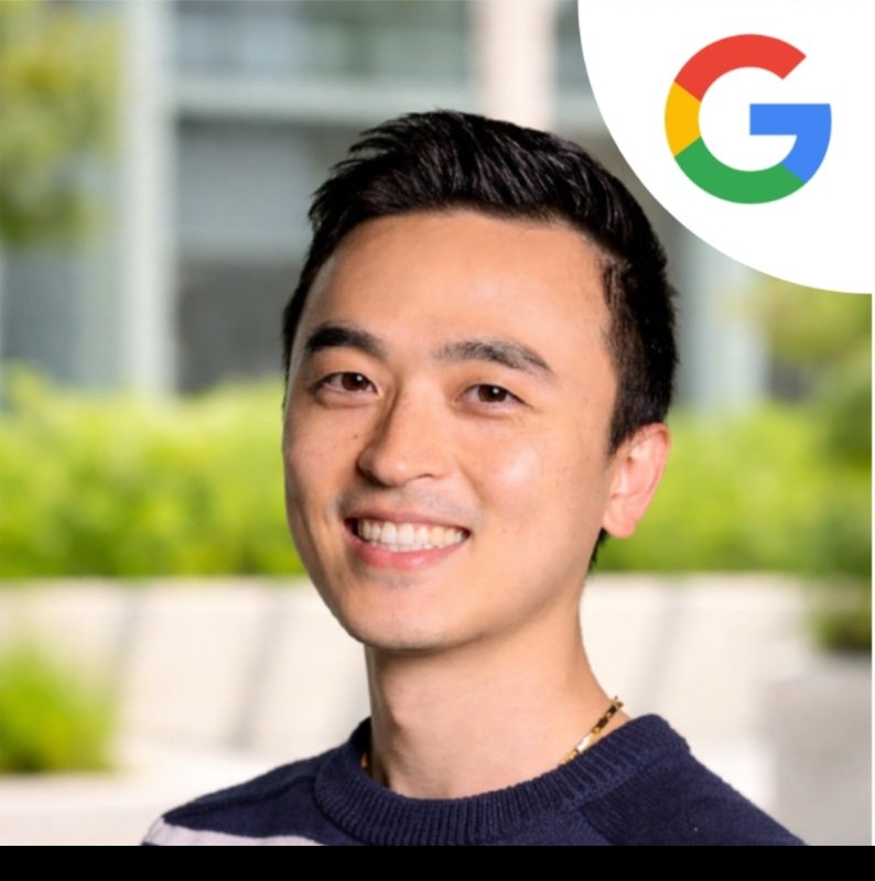 A smiling young man with short black hair wearing a navy sweater standing outdoors, with a blurred background of greenery and buildings, and a Google logo in the upper right corner.