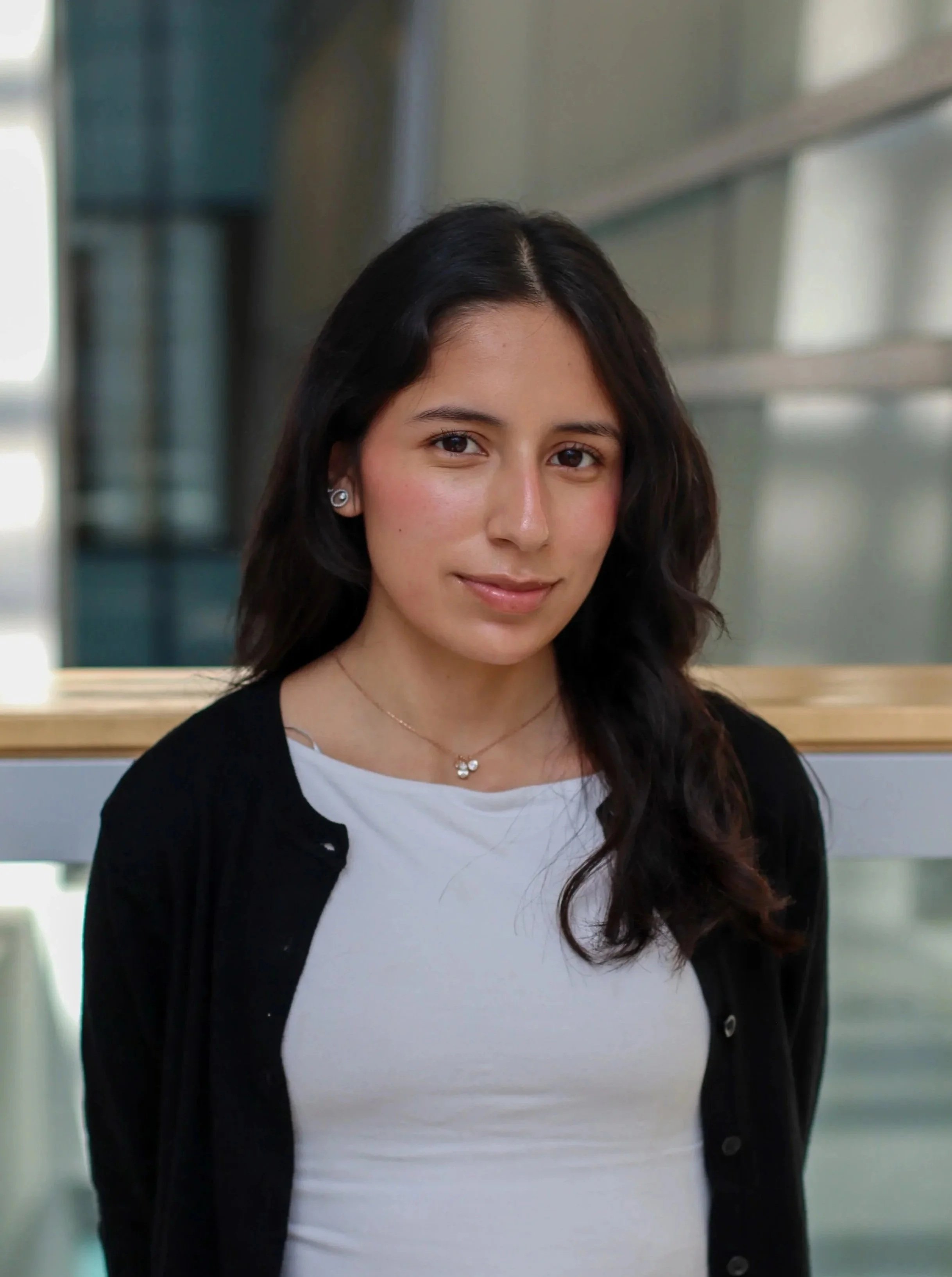 A young woman with long dark wavy hair wearing a white shirt, black cardigan, and a small necklace, standing in front of a modern interior with large windows.