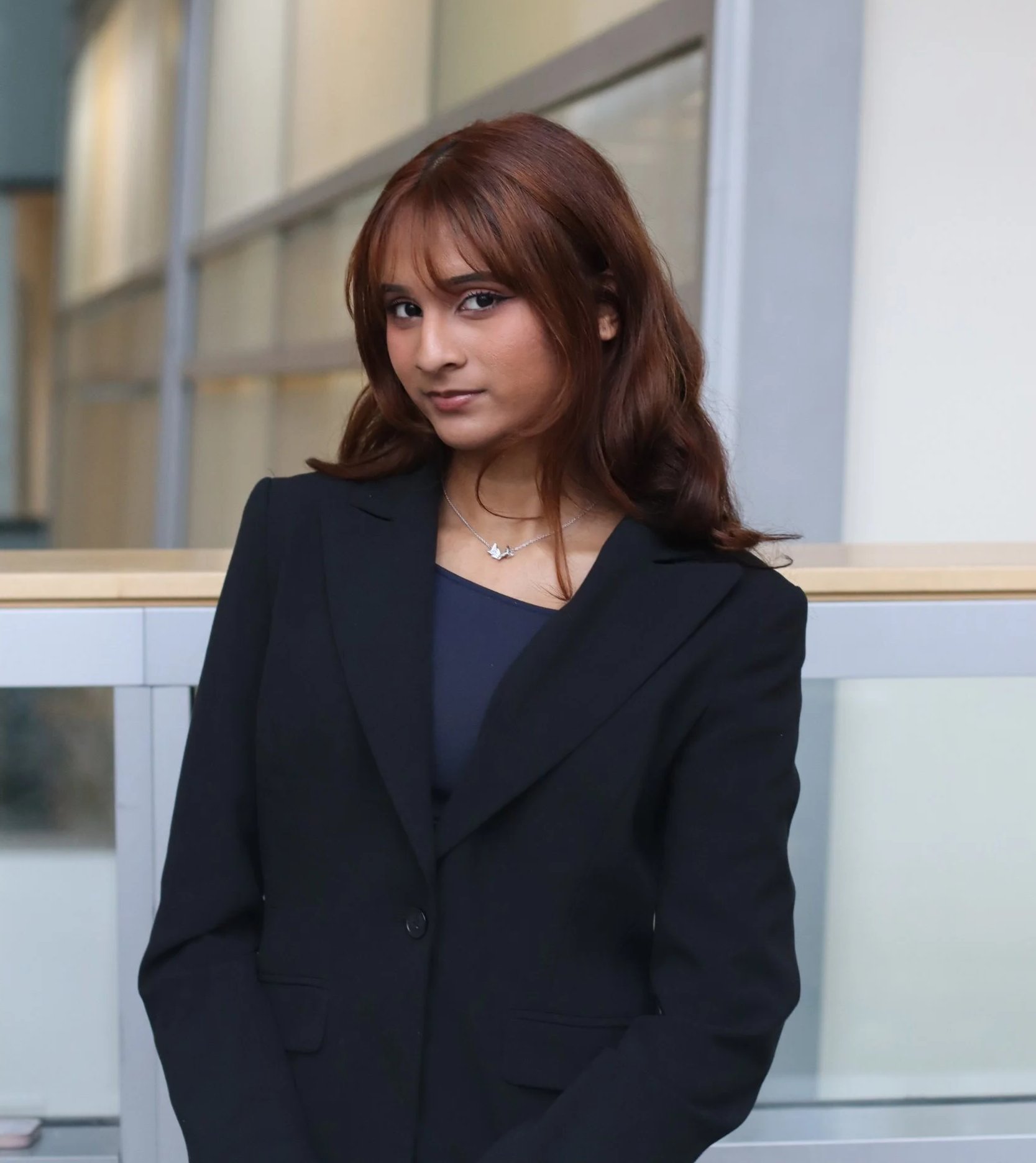 A woman with brown hair wearing a navy blazer and necklace, standing indoors against a background of a wall and glass partition.