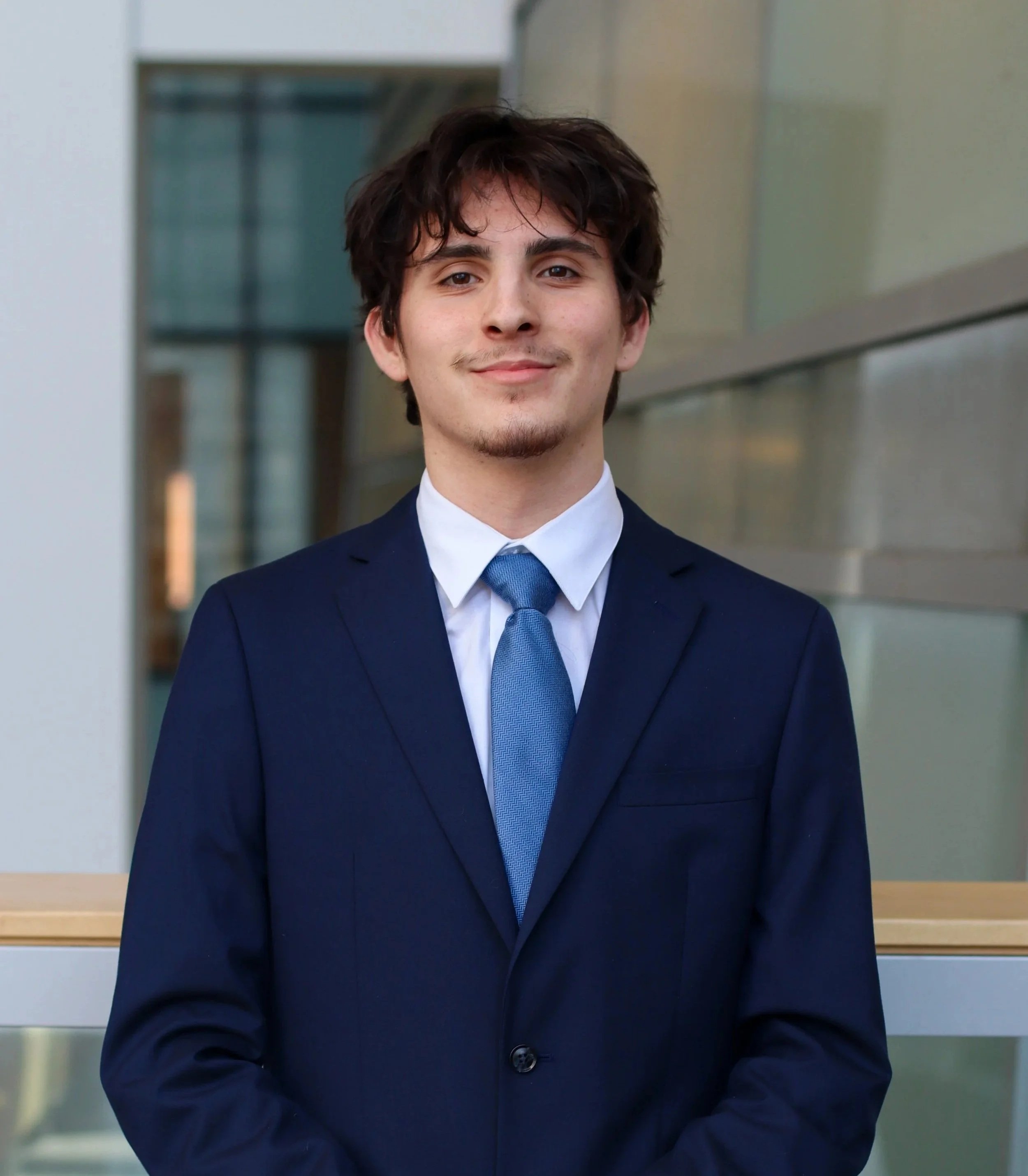 A young man with dark brown hair and a slight beard wearing a navy suit, white dress shirt, and a blue tie, standing outside with a modern building in the background.