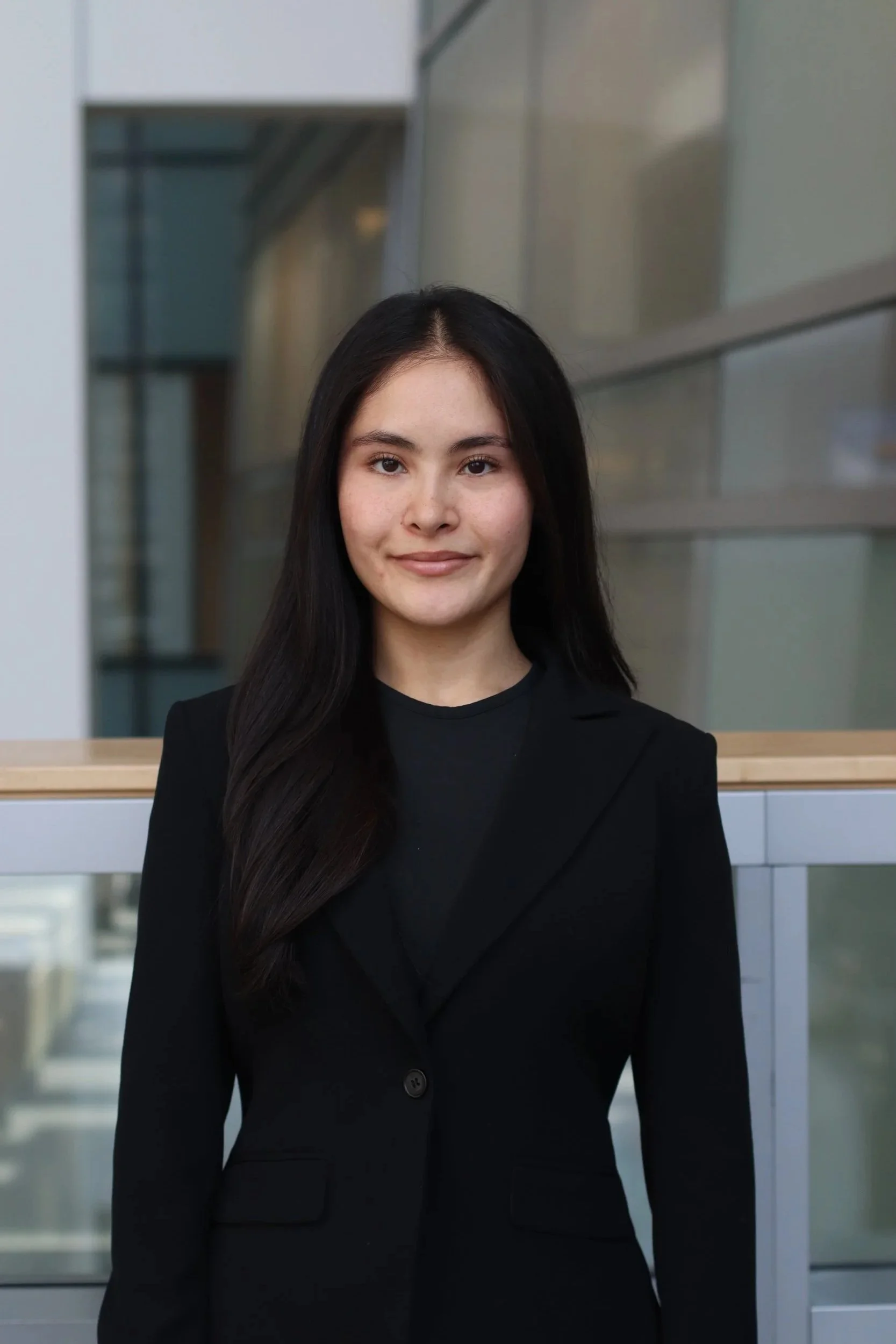 A young woman with long dark hair, wearing a black blazer and black top, standing in an indoor modern office or building with glass walls.
