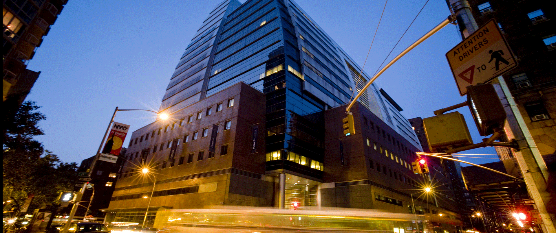 Tall modern glass building in New York City at dusk with streetlights, traffic lights, and street signs