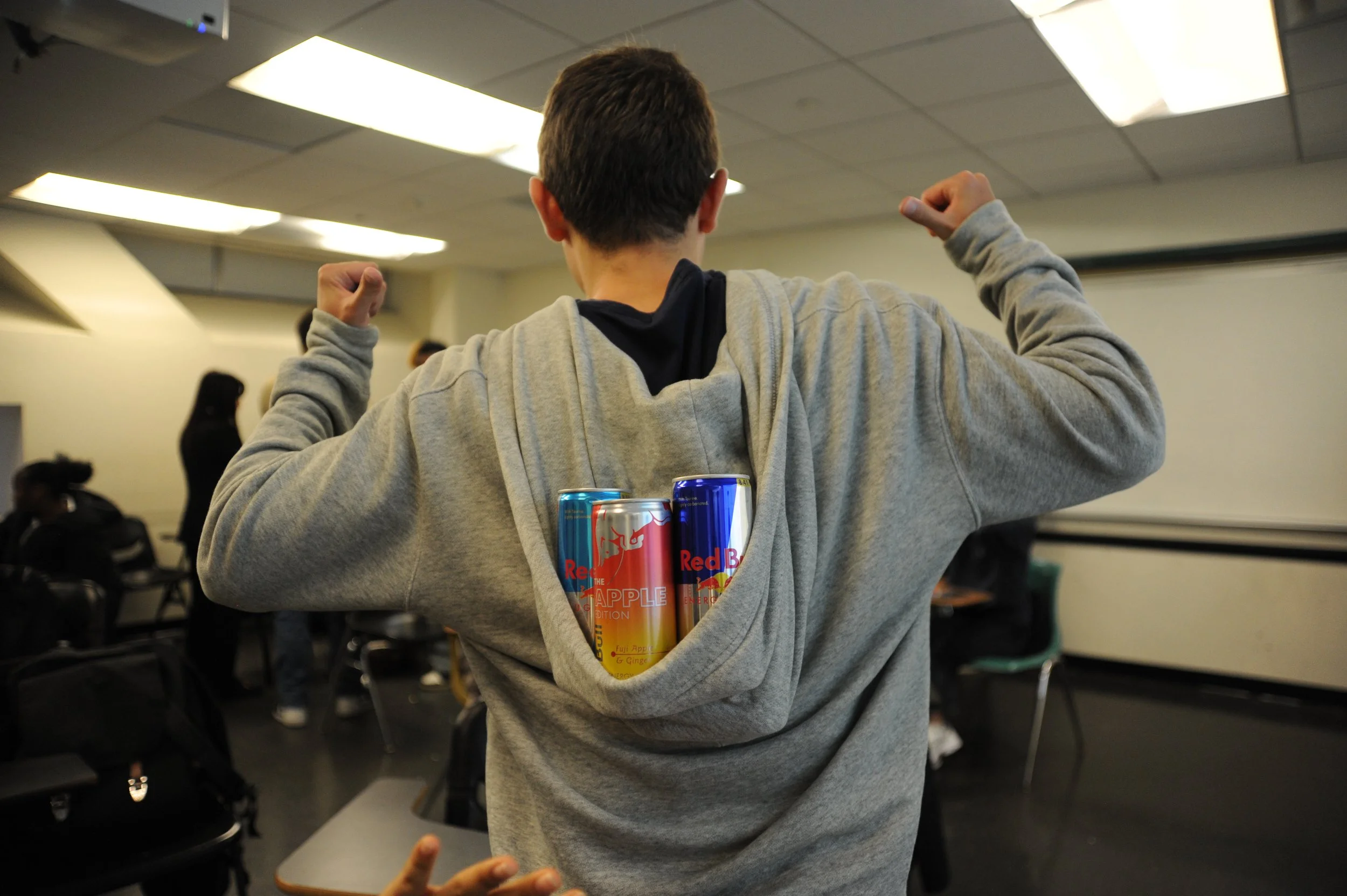 Person in a gray hoodie flexing with Red Bull energy drink cans tucked into the back of the hoodie