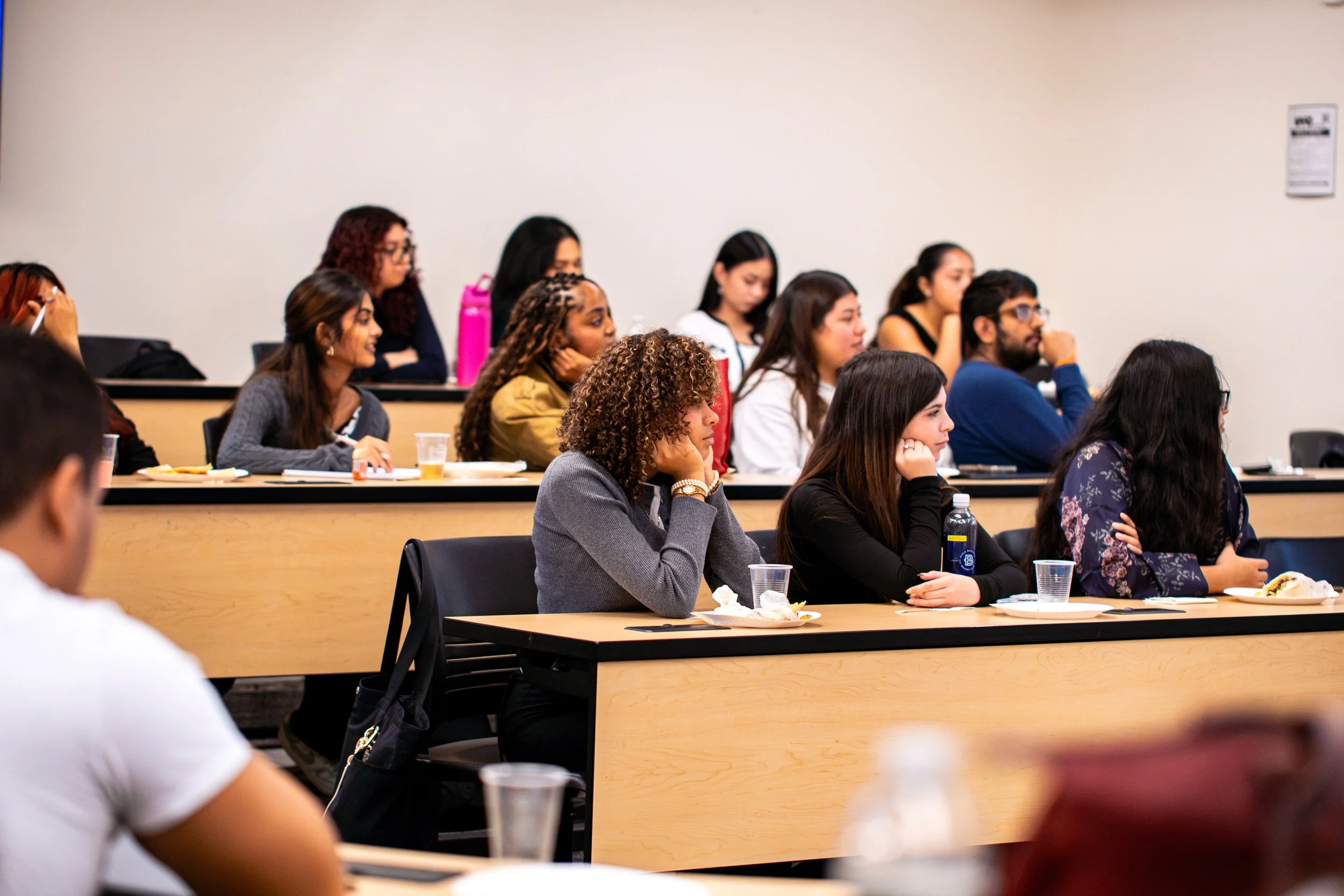 A diverse group of people attending a lecture or conference in a classroom setting, sitting at tables with food and drinks.