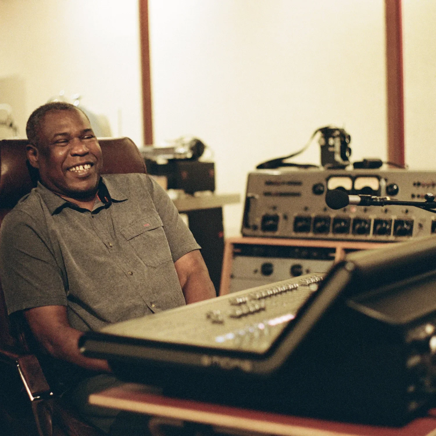 A man smiling and sitting in a recording studio with musical equipment around him, including a keyboard and a microphone.