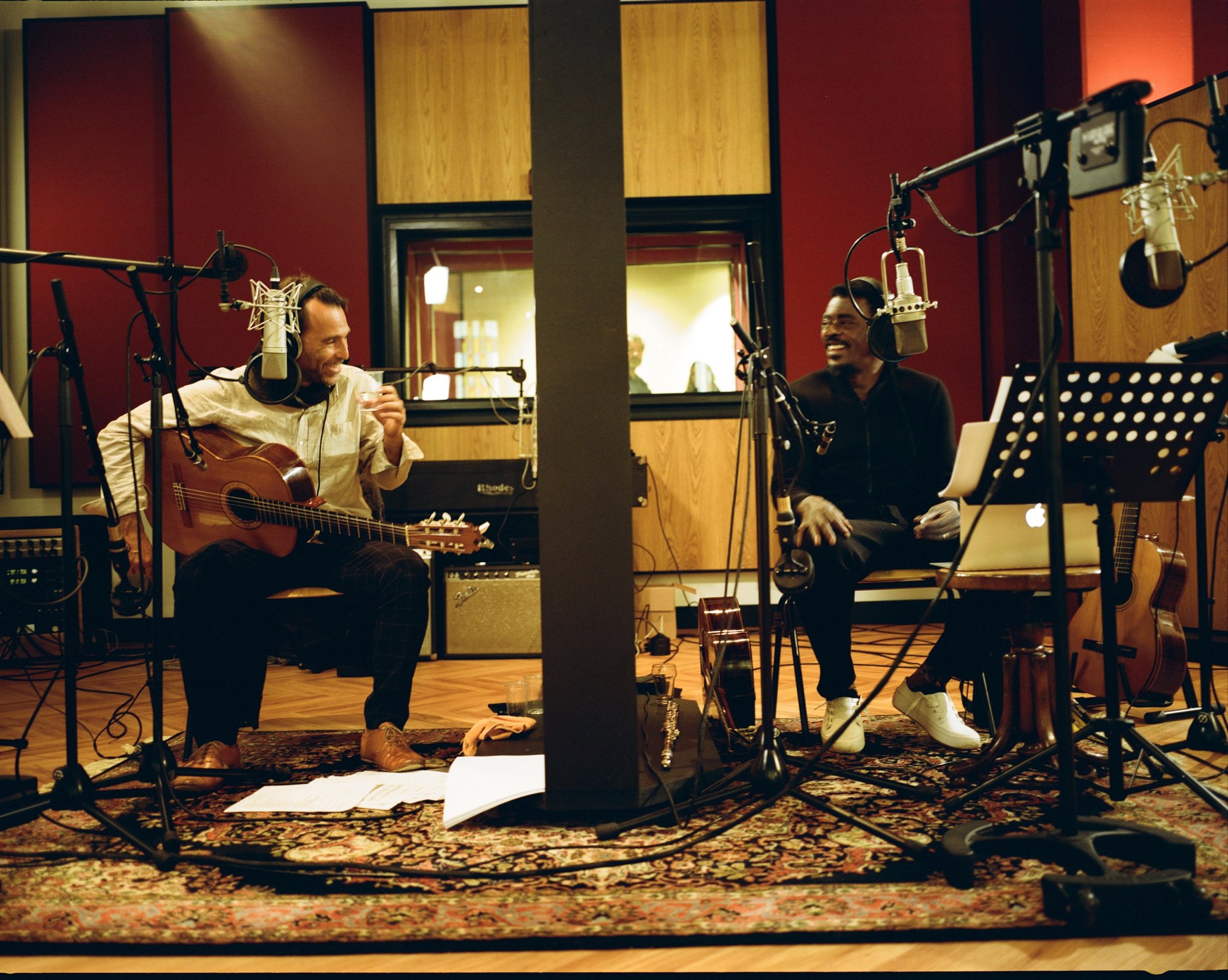 Two men in a recording studio sitting on chairs with microphones, talking and smiling, surrounded by musical instruments and music equipment.