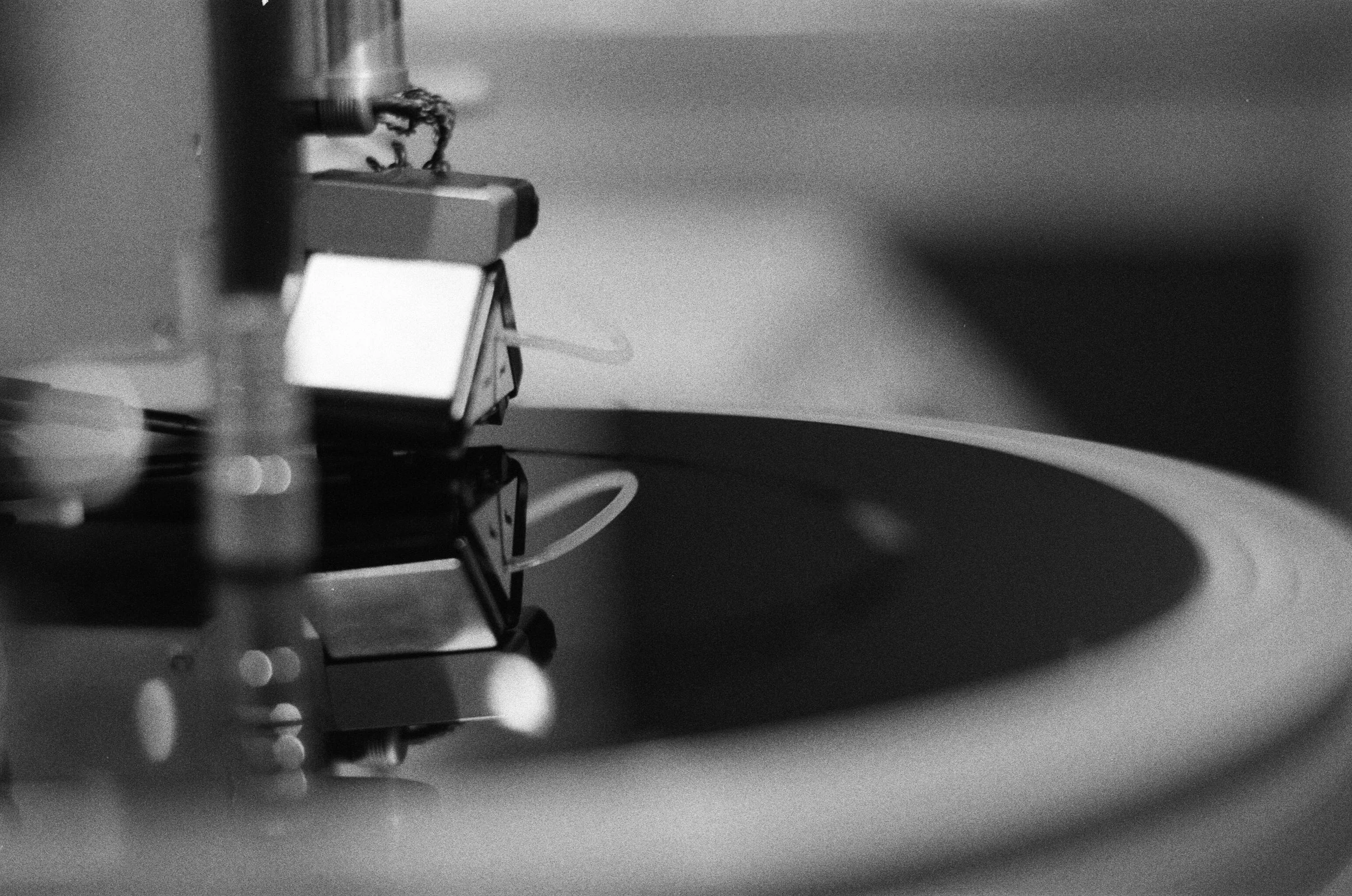 Close-up black-and-white photo of a turntable with a tonearm and stylus, spinning a vinyl record.