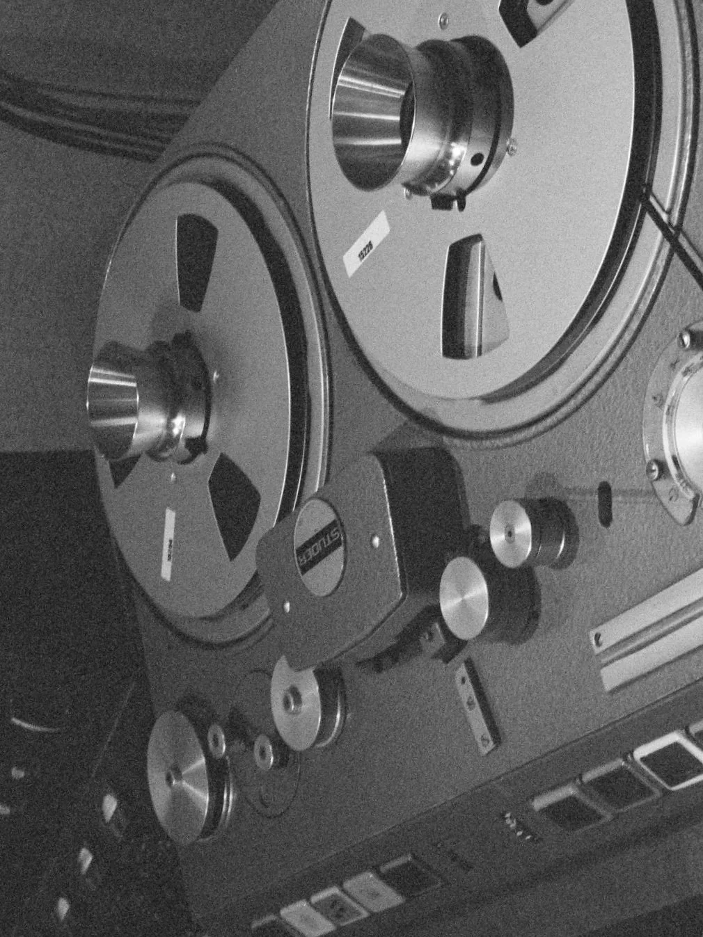 A vintage reel-to-reel tape recorder with two large spools, knobs, and control buttons on a grey panel.
