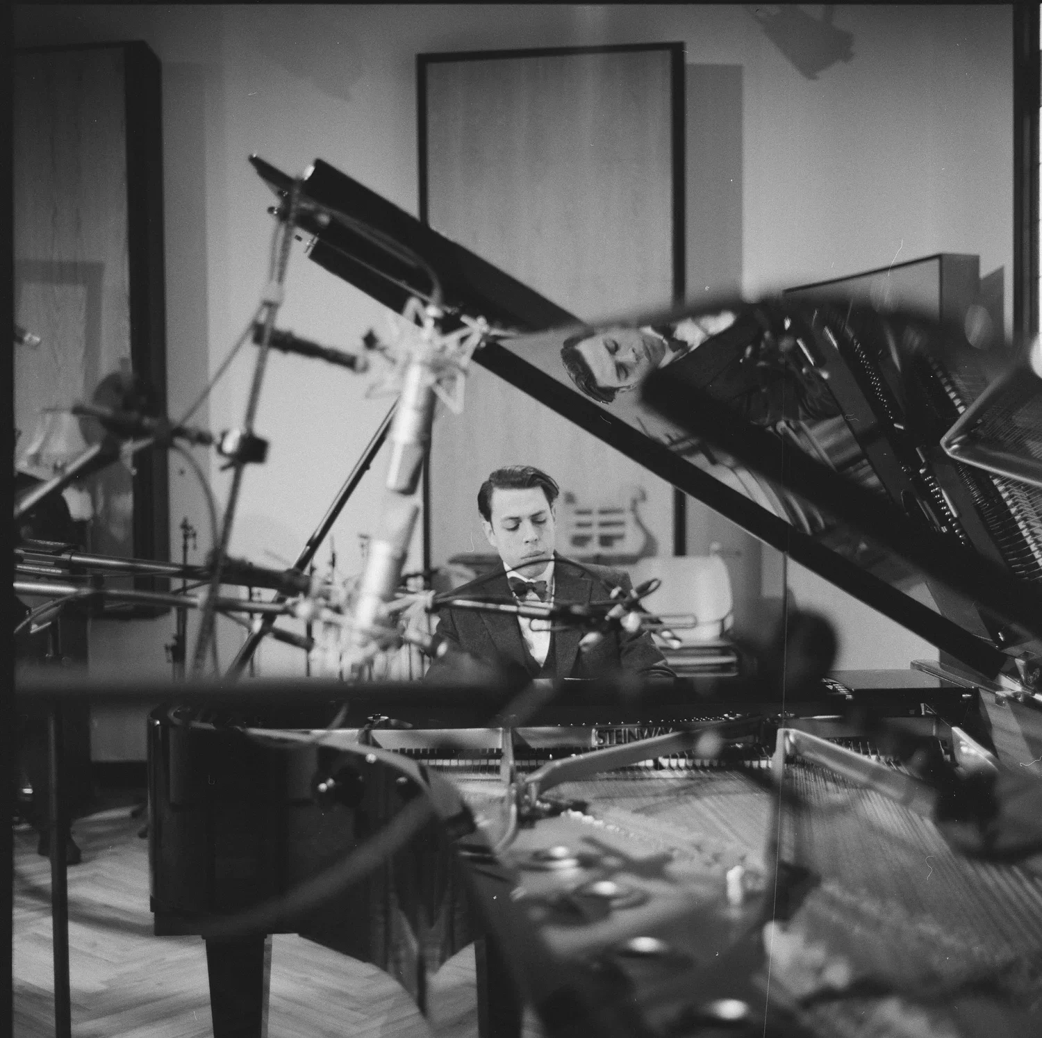 Black and white photo of a young man playing a grand piano in a music studio or performance space, with music stands and other equipment visible around him.