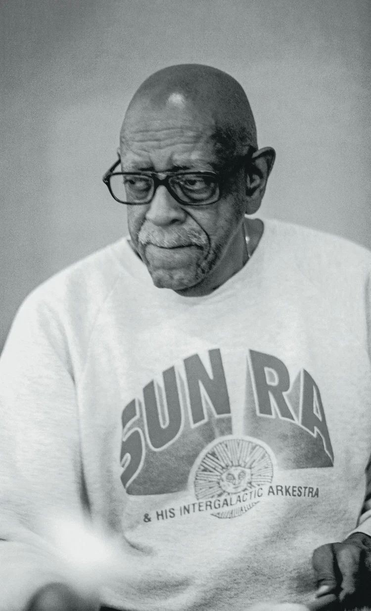A black and white photo of an elderly man with glasses, a bald head, and a white mustache and beard, wearing a T-shirt with the words 'SUN RA & HIS INTERGALACTIC ARKESTRA' and a graphic of a sun with a face.