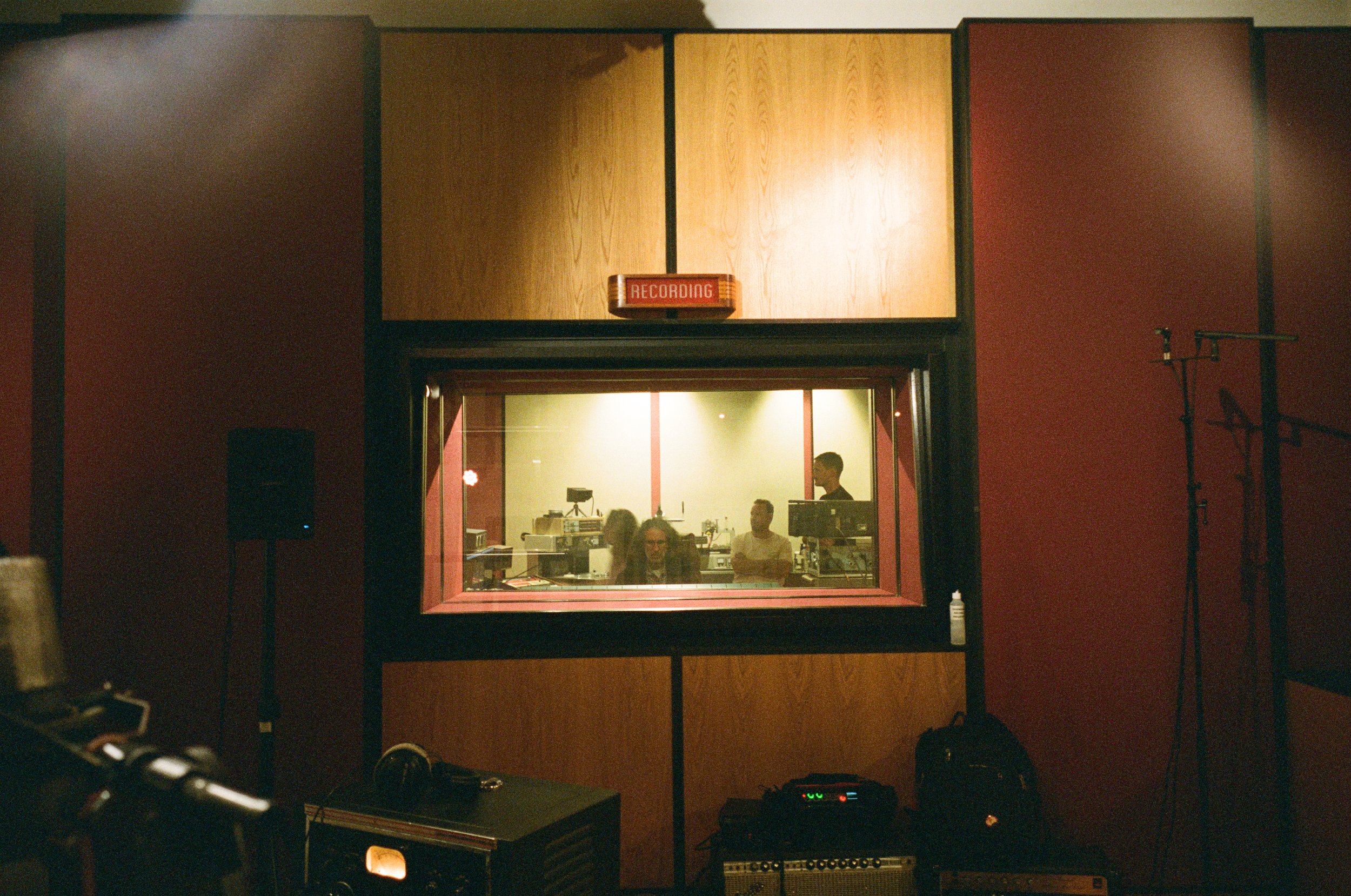 A recording studio control room viewed through a window with people working inside, machines, and audio equipment visible.