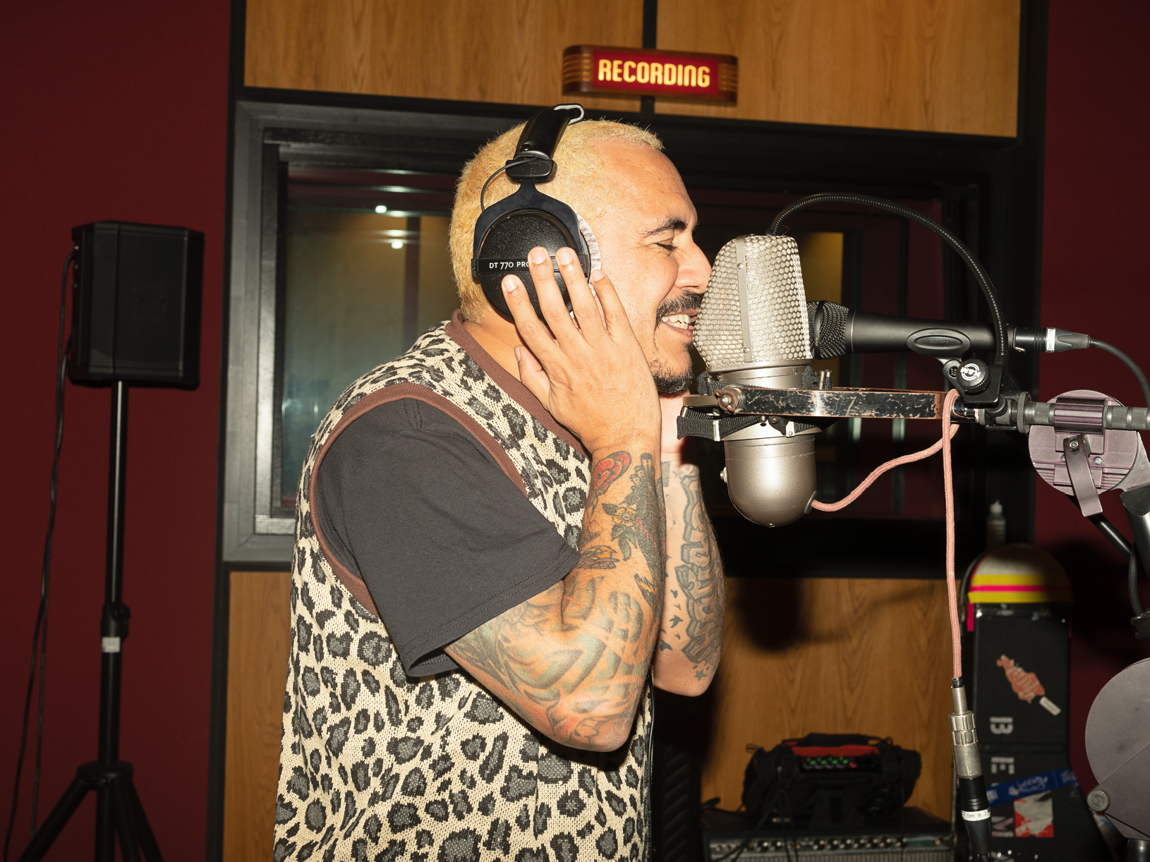 A man with tattoos and blonde hair wearing a leopard print vest over a black shirt singing into a large microphone in a recording studio, holding headphones to his ear.