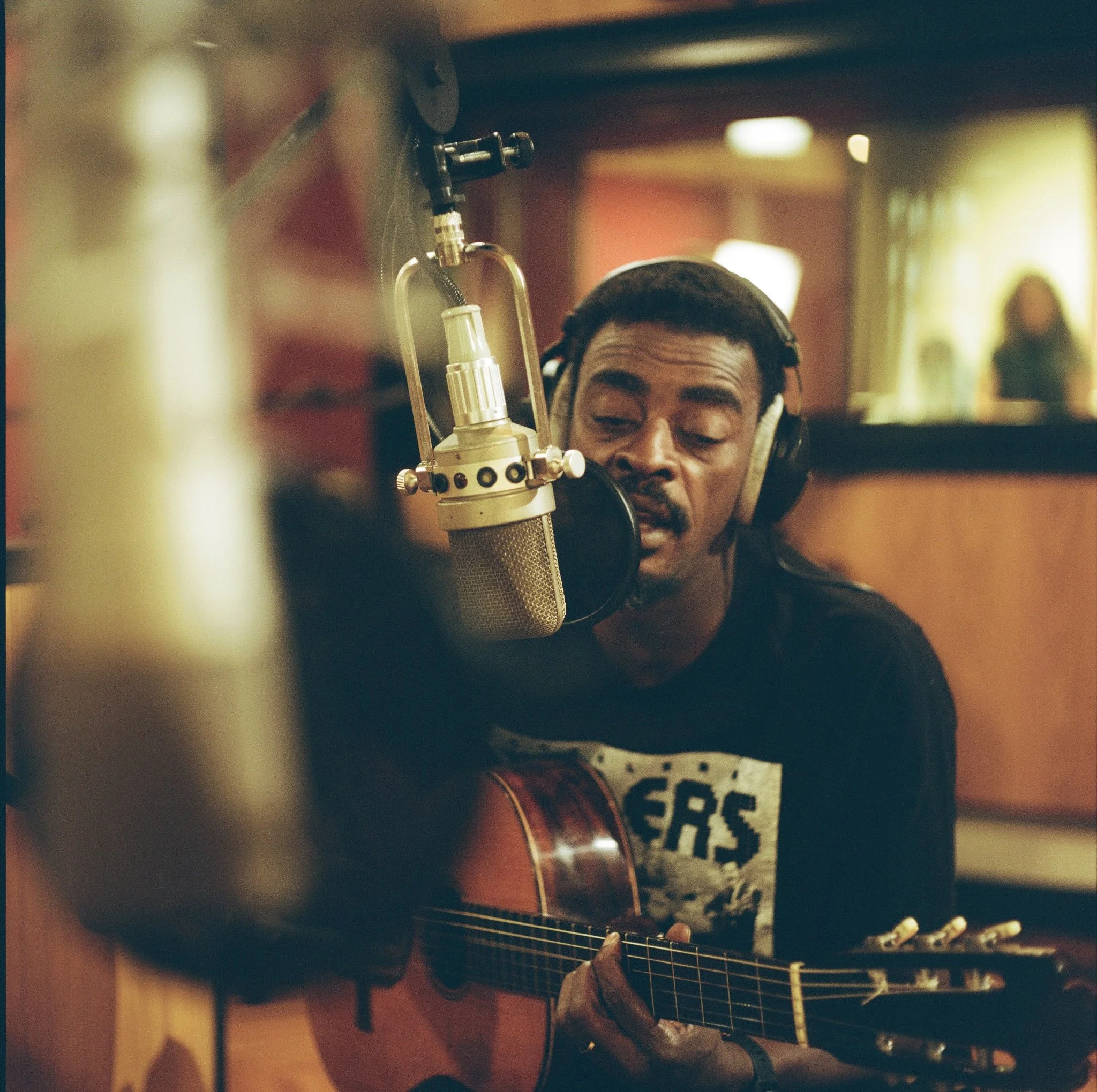 A man with dark hair and a mustache, wearing headphones, playing an acoustic guitar in a warmly lit room, with a blurred woman in the background.