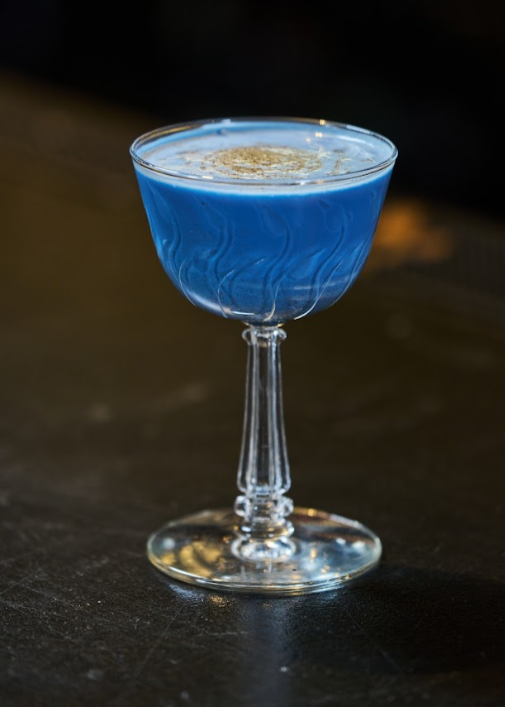 A fancy cocktail glass filled with bright blue liquid, garnished with a lemon twist, on a dark wooden surface.