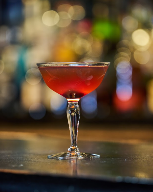 A cocktail in a classic coupe glass with a red drink inside, set on a bar counter with a blurred colorful background.