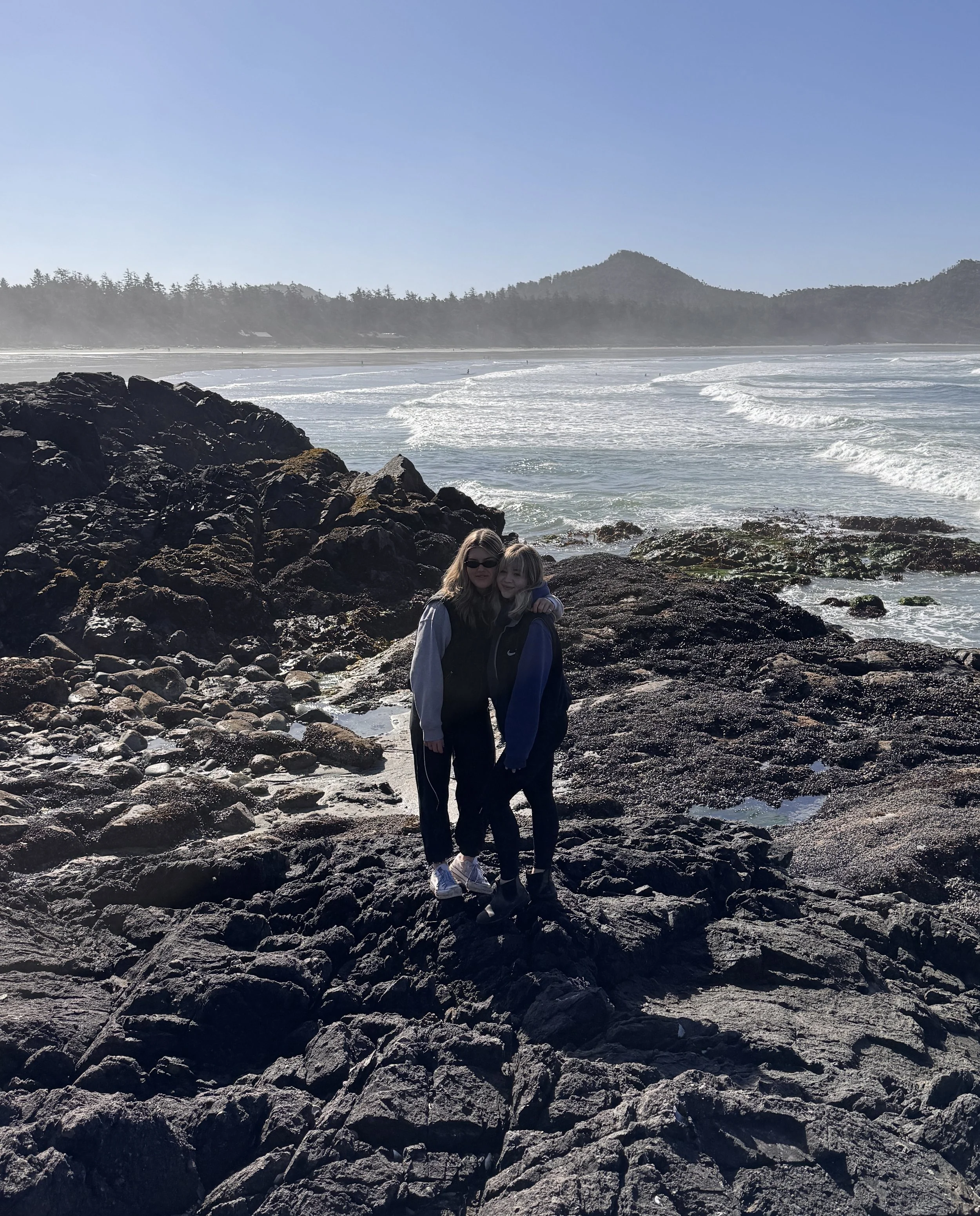 Two women standing on a rocky shoreline near the ocean, with waves and a distant tree-lined coast and hills in the background.