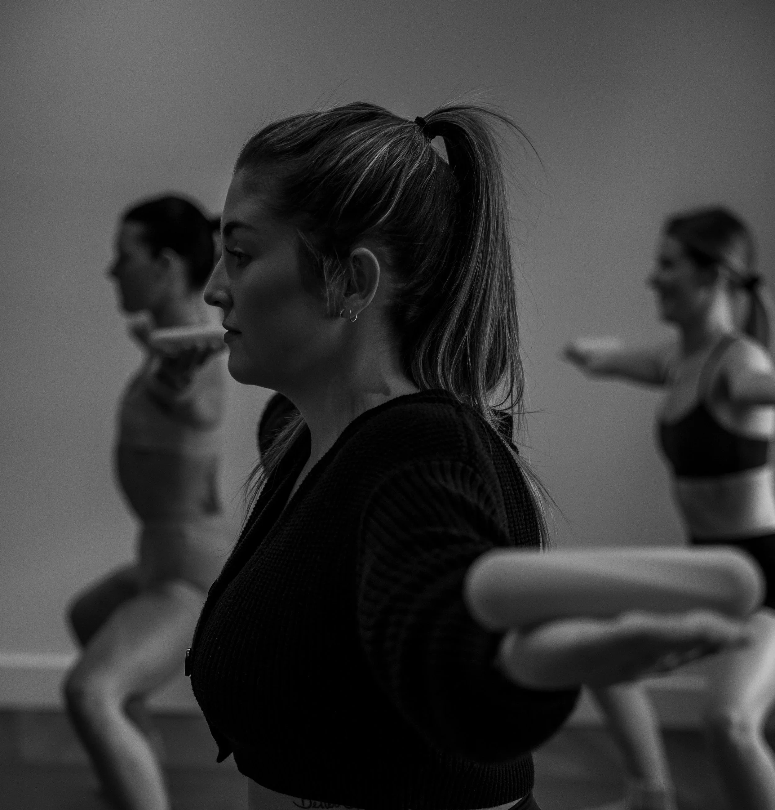 A black-and-white photo of three women with braided hair standing side by side, facing downward, in what appears to be a yoga or meditation class.