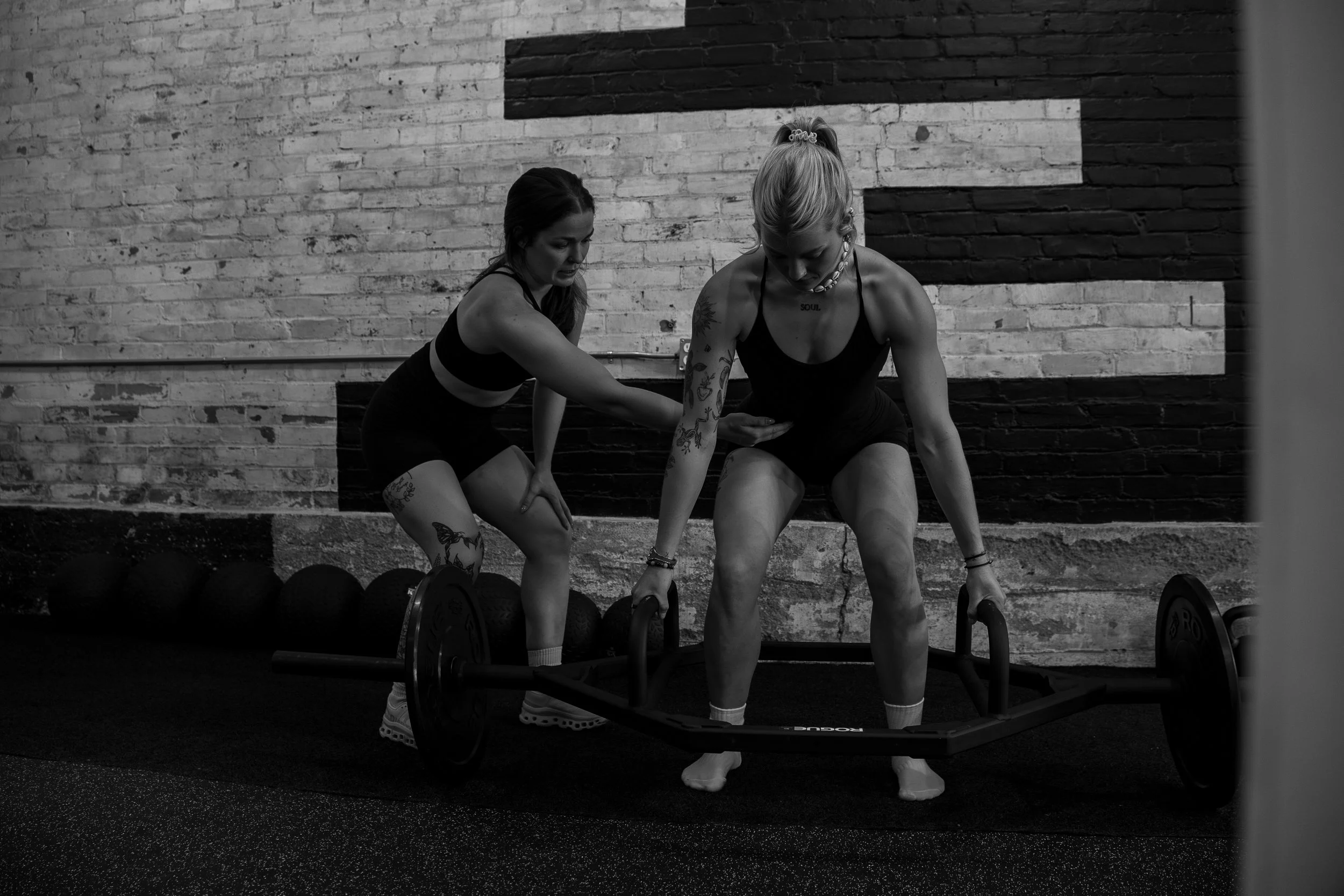 Two women using kettlebells during a workout in a gym with a brick wall background.