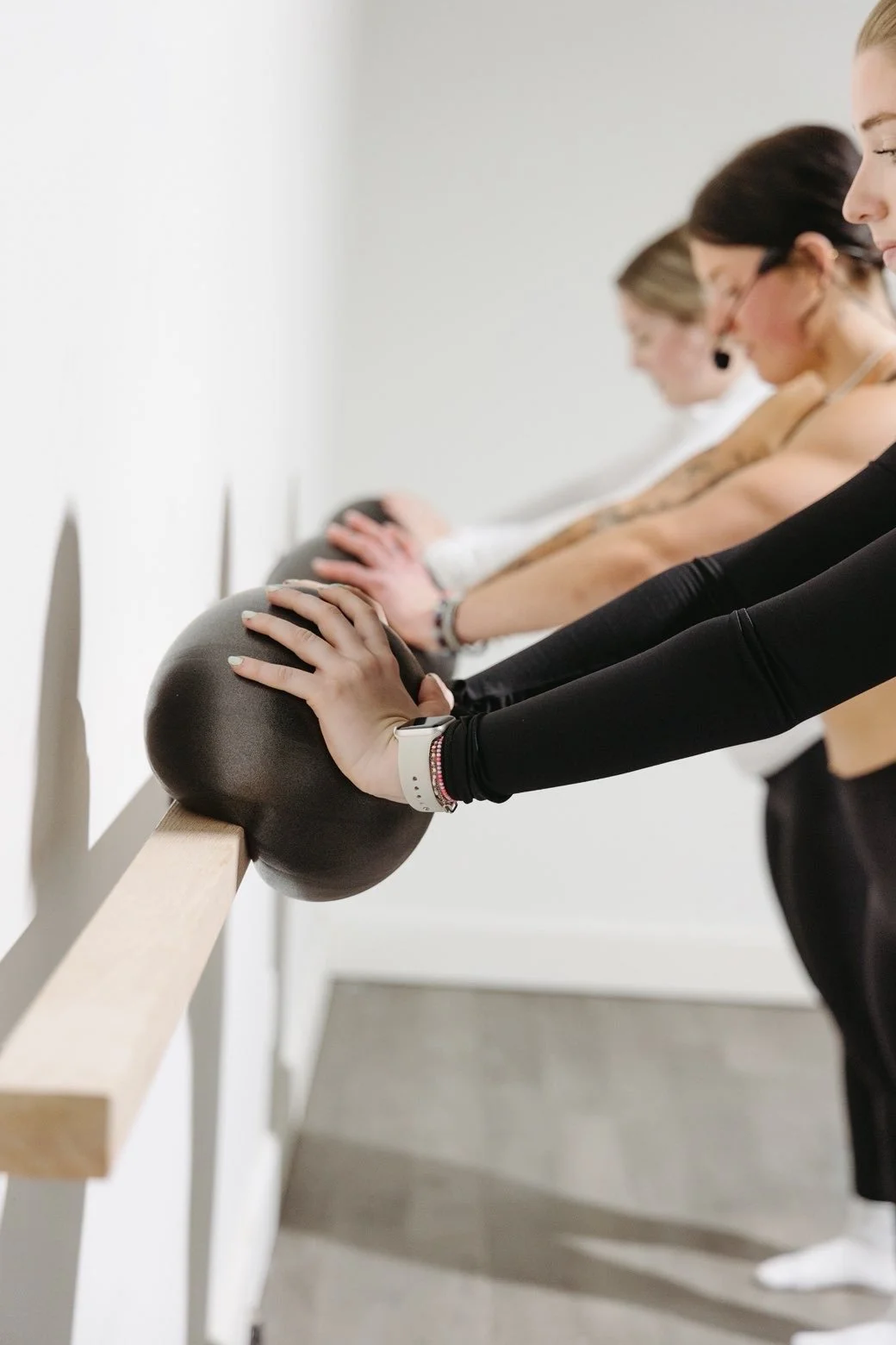 Four women are doing a stretching exercise with their hands on exercise balls, leaning forward against a wall in a fitness class.