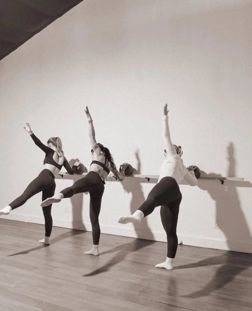 Three women in activewear doing Barre Pilates in a studio, balancing on one leg with arms raised, with a ballet bar and slippers on the wall behind them.