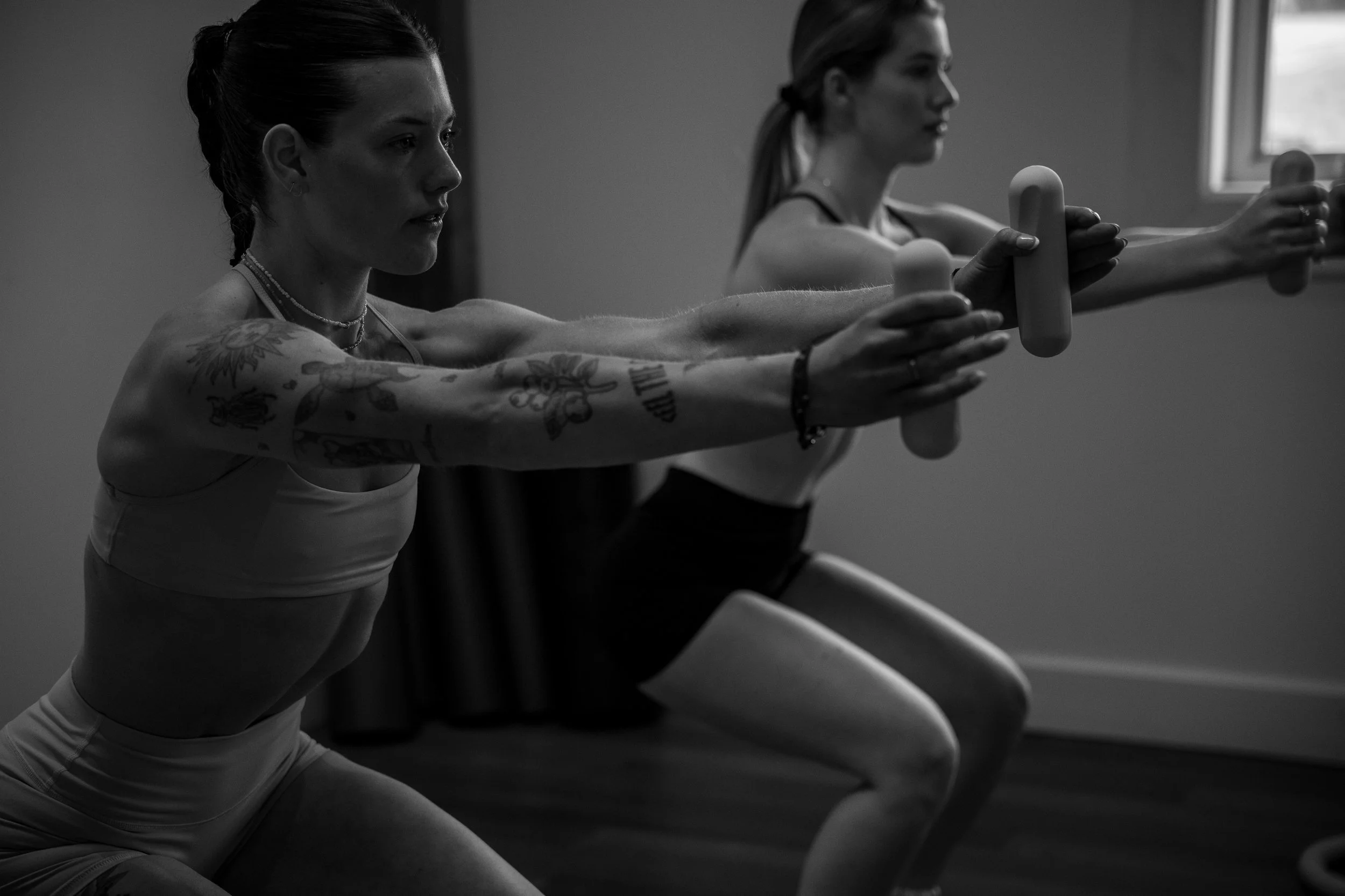 Two women in workout clothes stretching with resistance bars during a fitness class in a room with natural light.