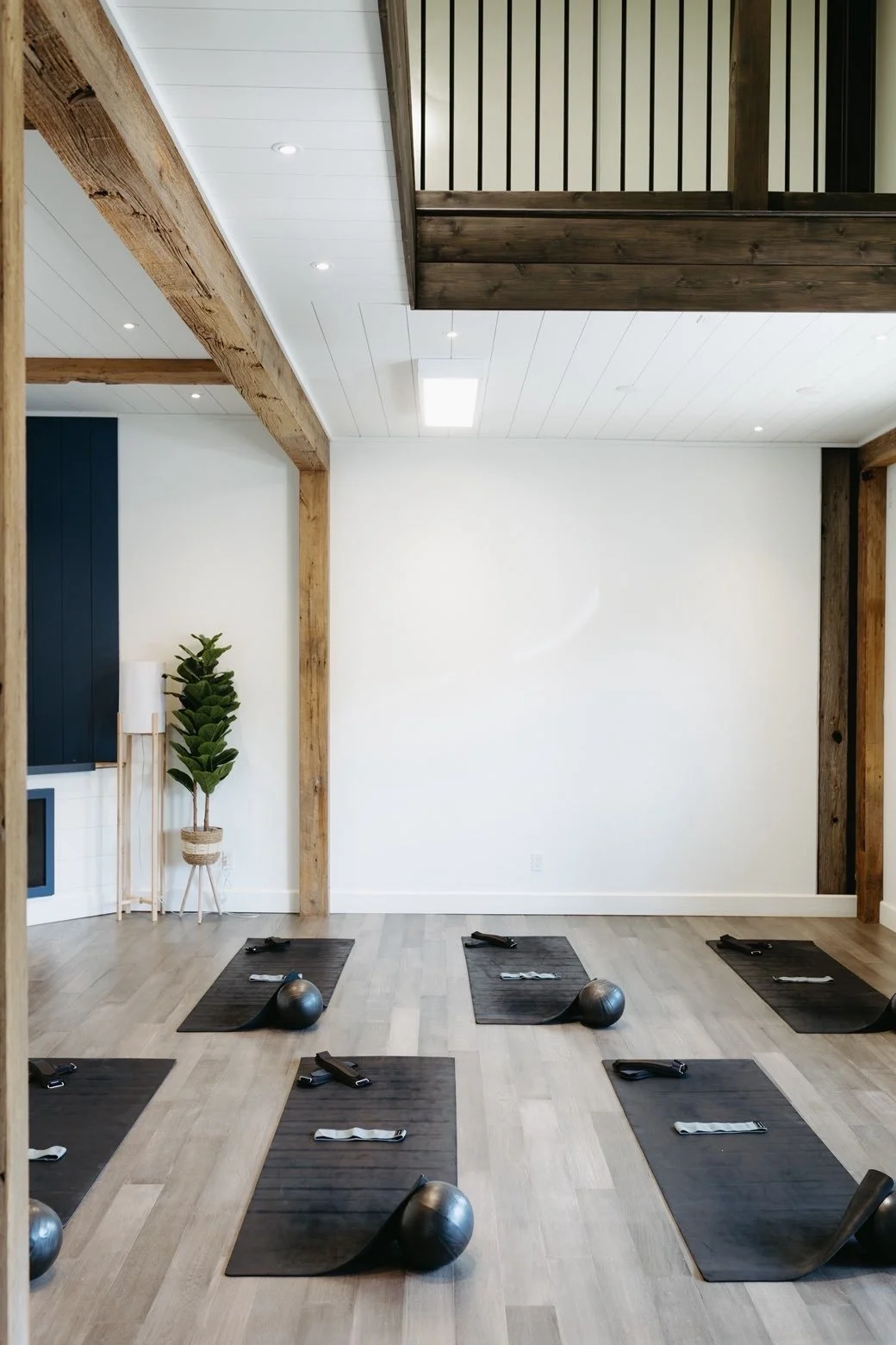 Interior of a yoga or fitness studio with black mats, exercise balls, and resistance bands arranged on a light wood floor. The space features a white wall, wooden beams, and a small elevated area with a staircase.