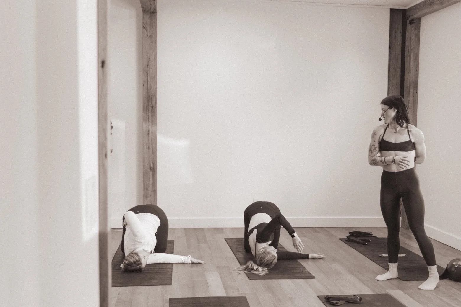 A yoga instructor with tattoos and dark hair leading a yoga class in a wooden-floored studio, with two women in child's pose on mats. The instructor is standing with a microphone headset, observing the students.