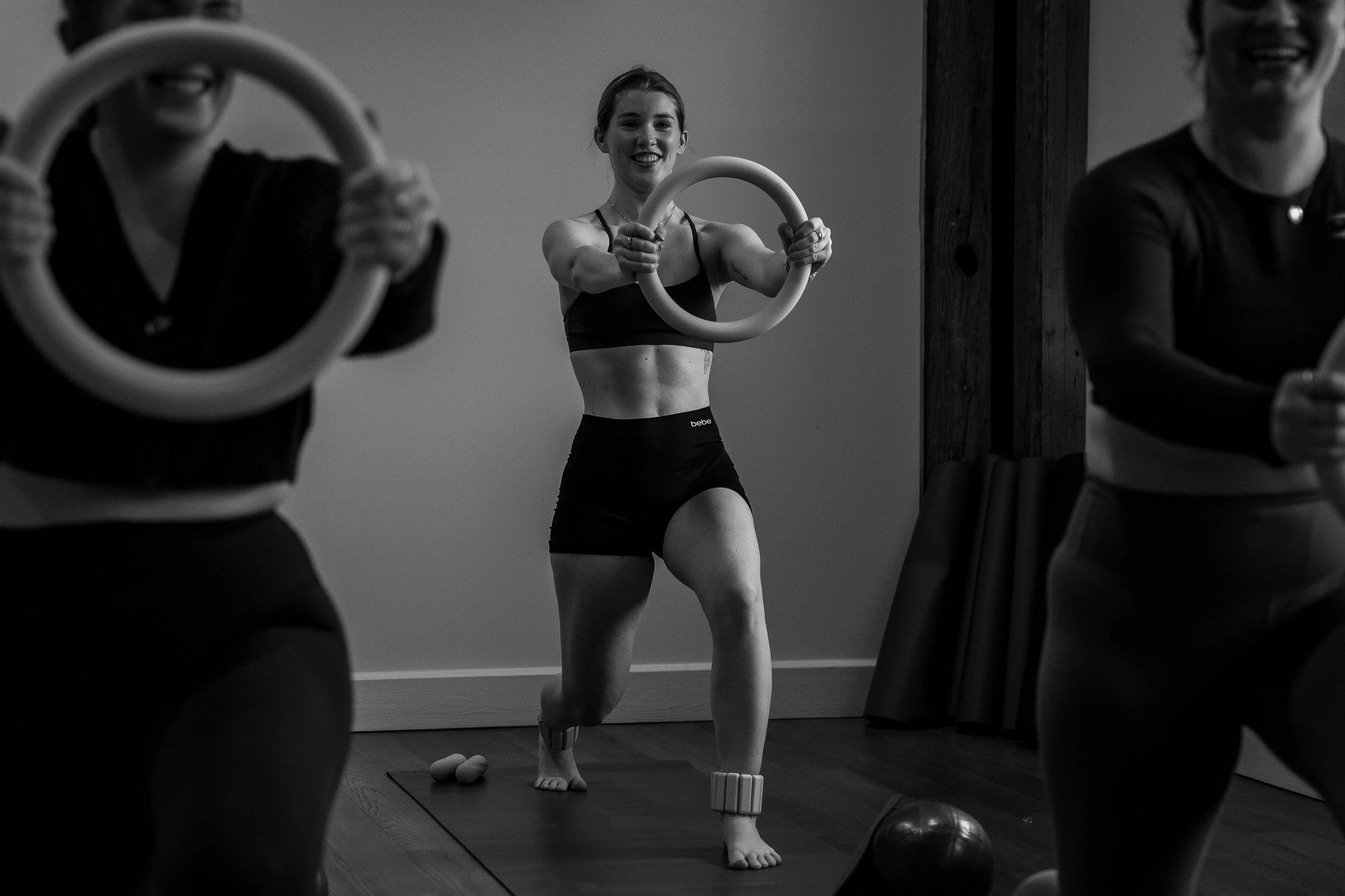 A woman performs a fitness exercise with a ring, while two other women hold rings in a studio. The woman in the center smiles and looks at the mirror, wearing athletic clothing, and stands on one leg.