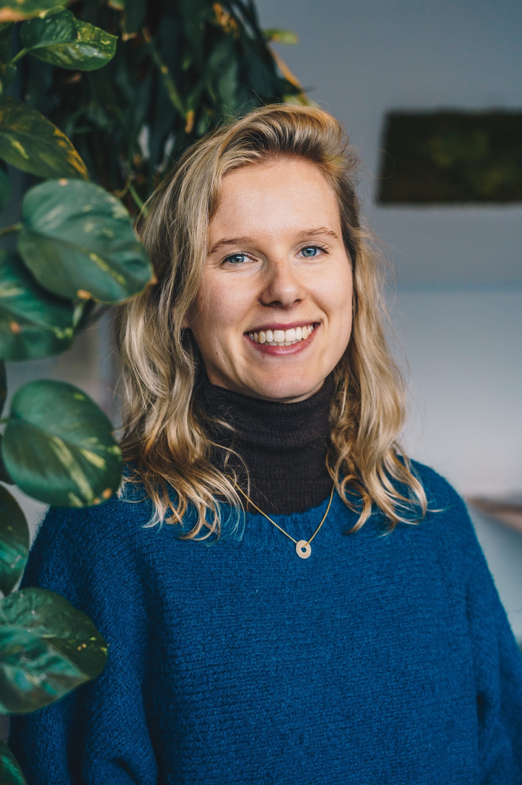 Smiling young woman with wavy blonde hair, wearing a black turtleneck and blue sweater, standing near a leafy green plant with a gray wall background.