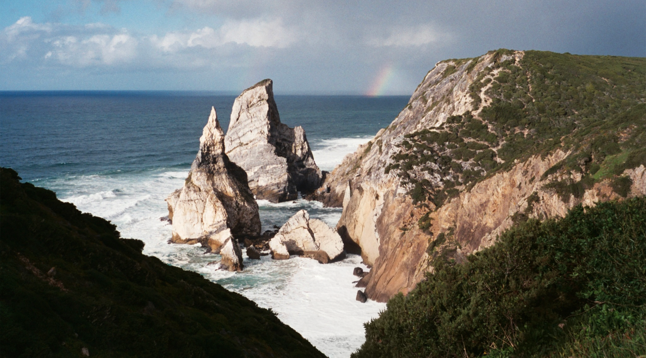 A coastal landscape featuring large white rock formations in the ocean near a green, rugged hillside with a faint rainbow in the background and a partly cloudy sky.