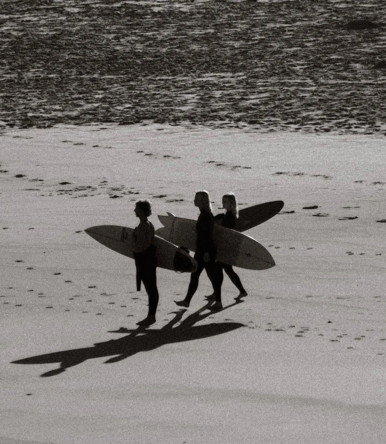 Three surfers walking on a beach, each holding a surfboard, with their shadows cast on the sand, in a black and white photo.