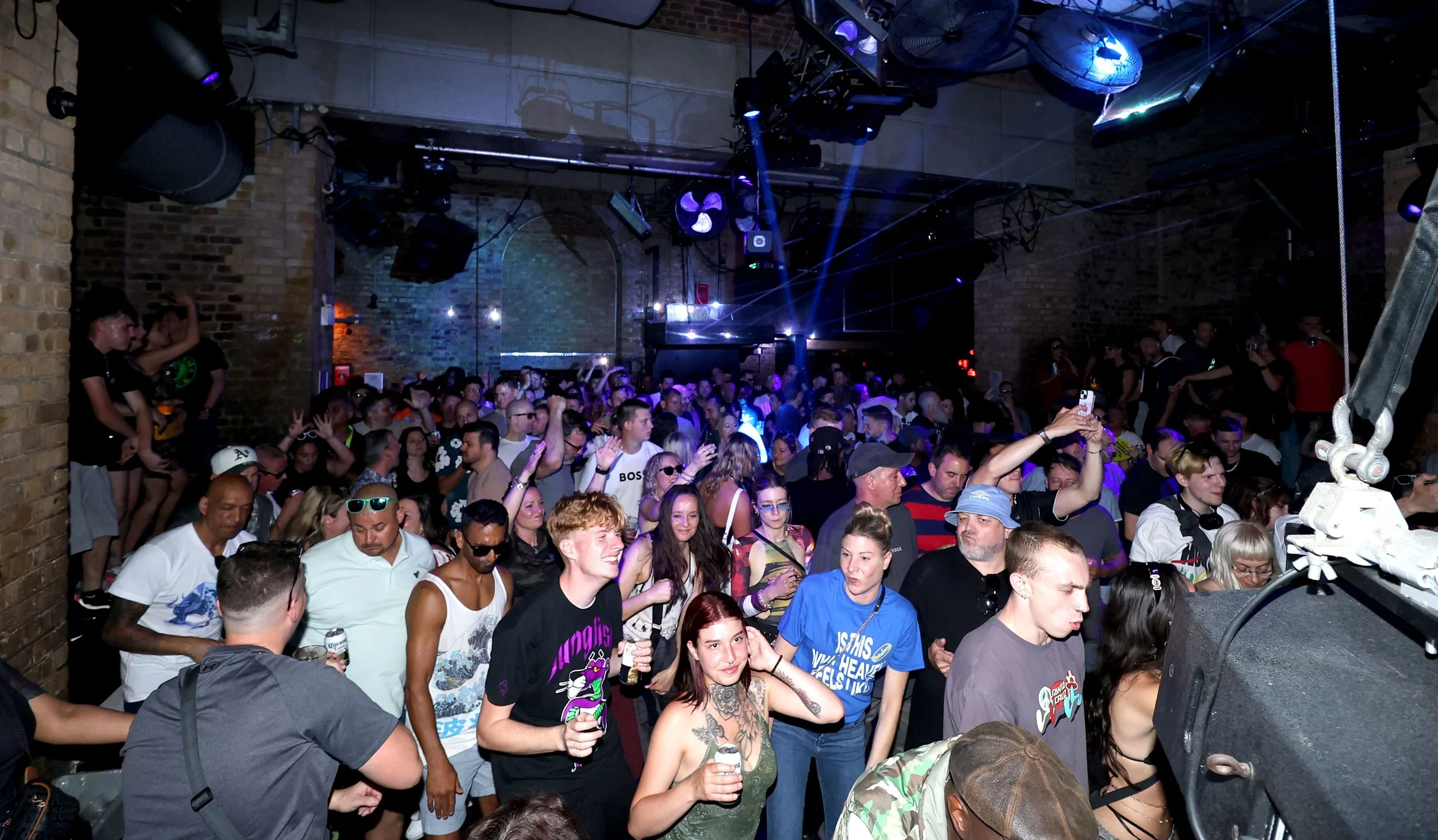 Crowd of people dancing and socializing in a nightclub with brick walls and blue lighting.