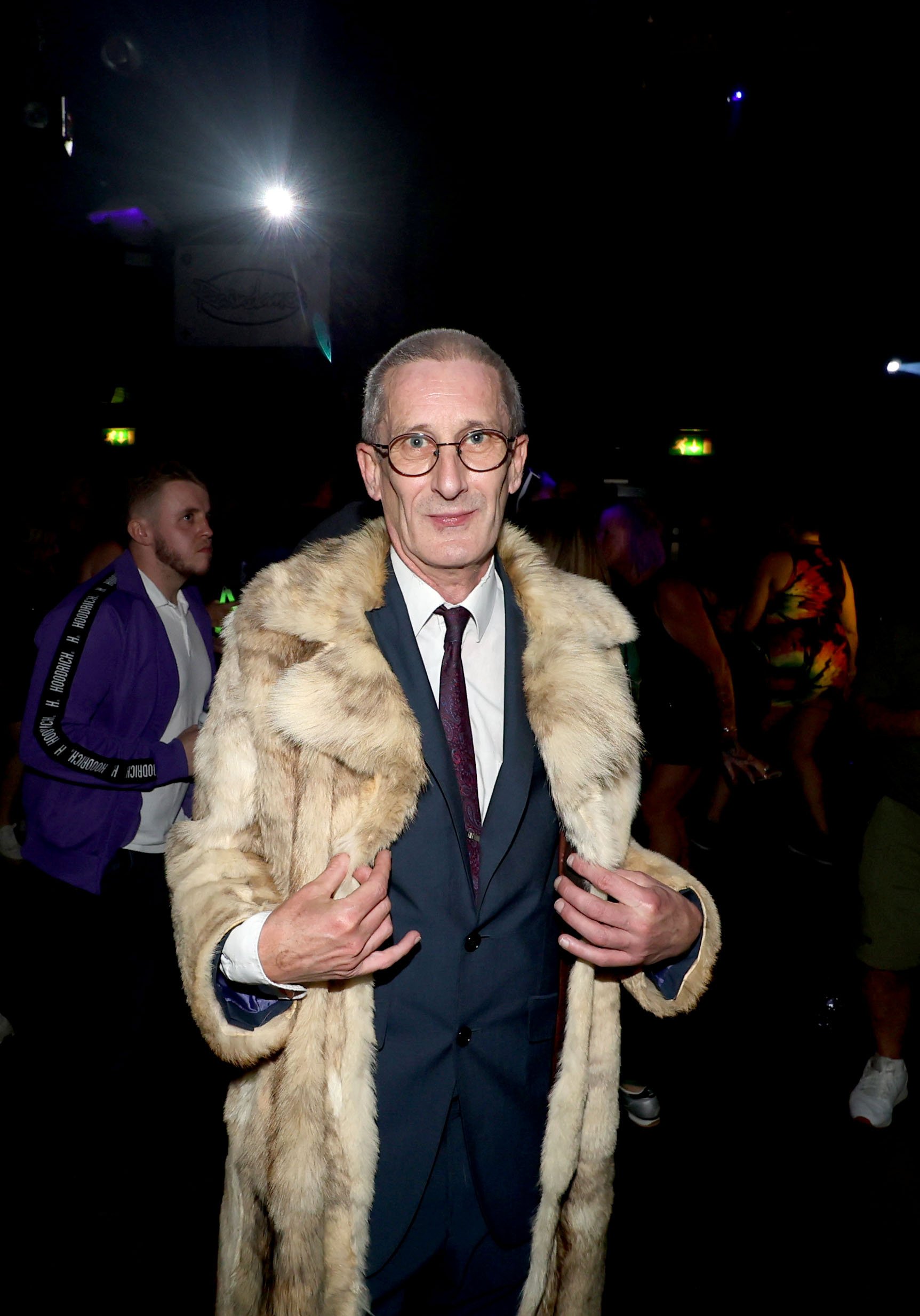 Man in a tuxedo and glasses wearing a fur coat at a party or nightclub, with other people in the background and dark lighting.