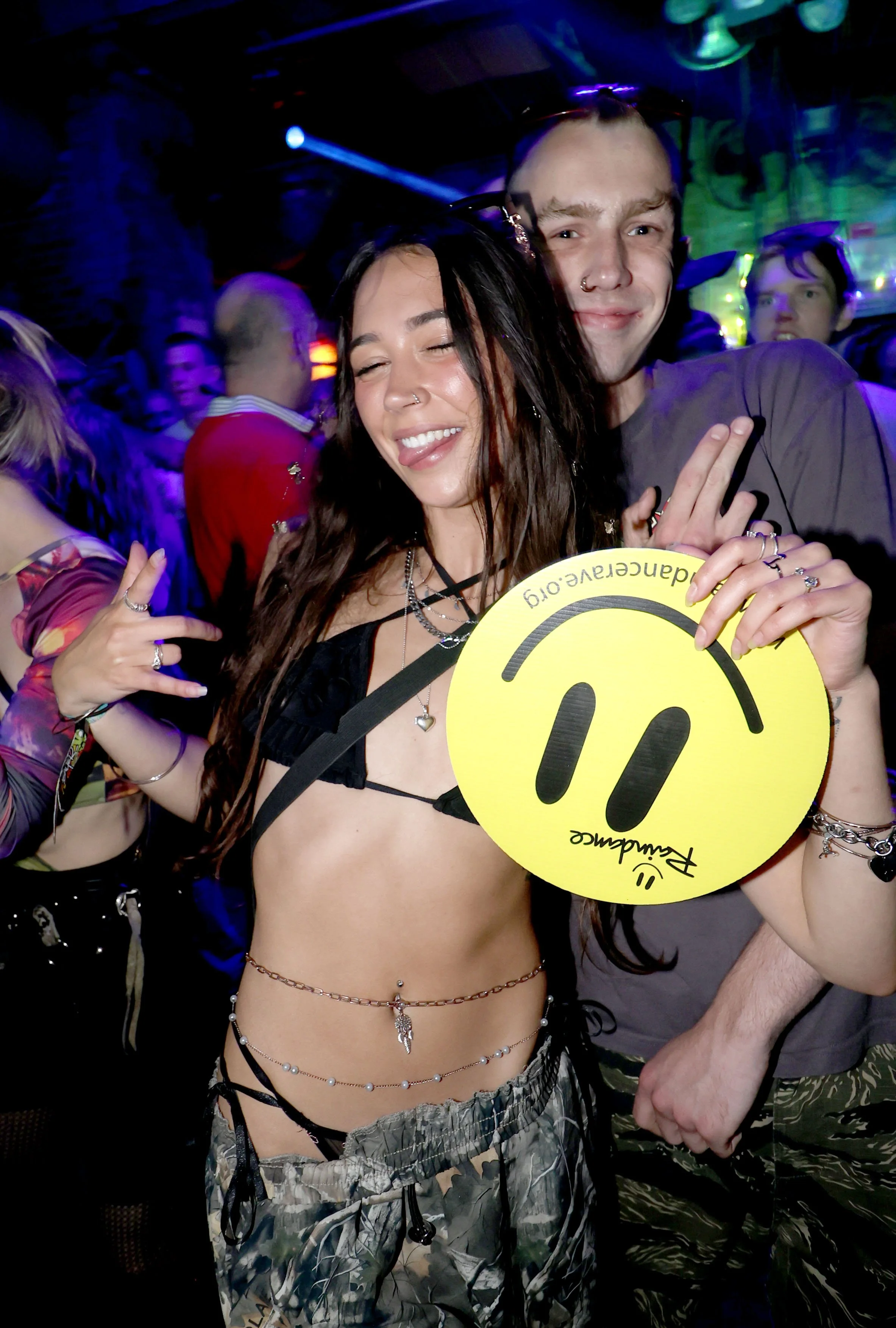 A woman with long dark hair smiling in a crowd at a party or concert, holding a yellow smiley face sign with headphones and the text 'raindancerave.org' and 'Raindance' written on it, wearing a revealing black top, camouflage pants, and jewellery.
