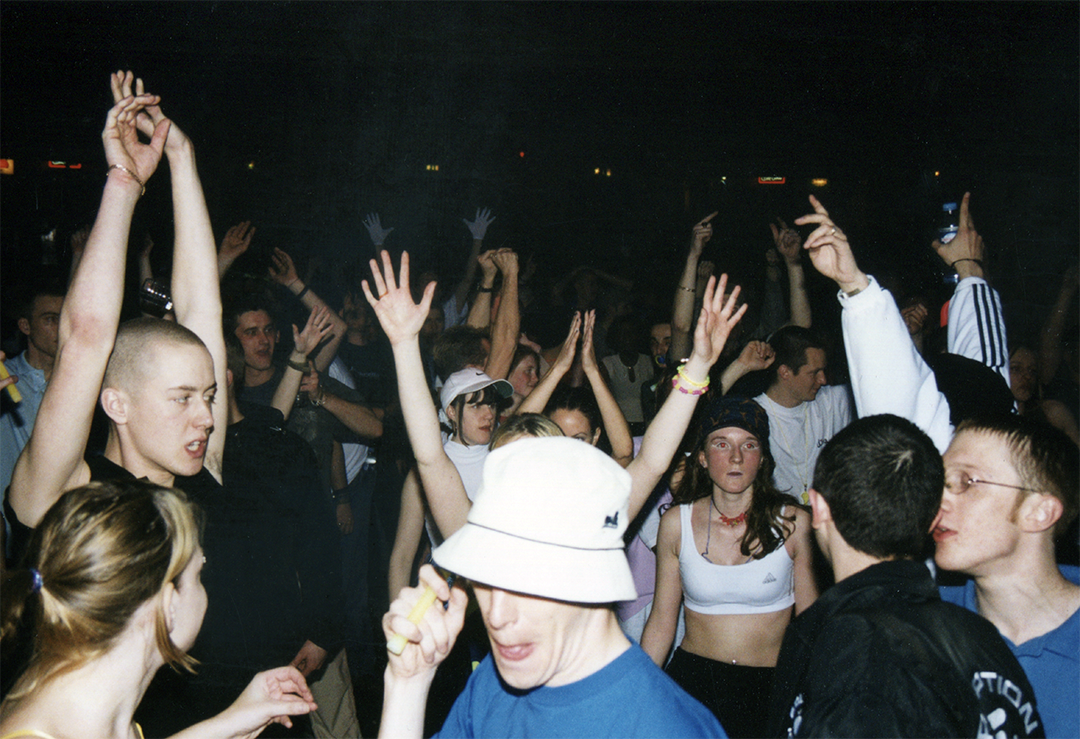 A group of young people dancing and having fun at a crowded indoor party or concert, with some raising their hands and others talking, under dim lighting.