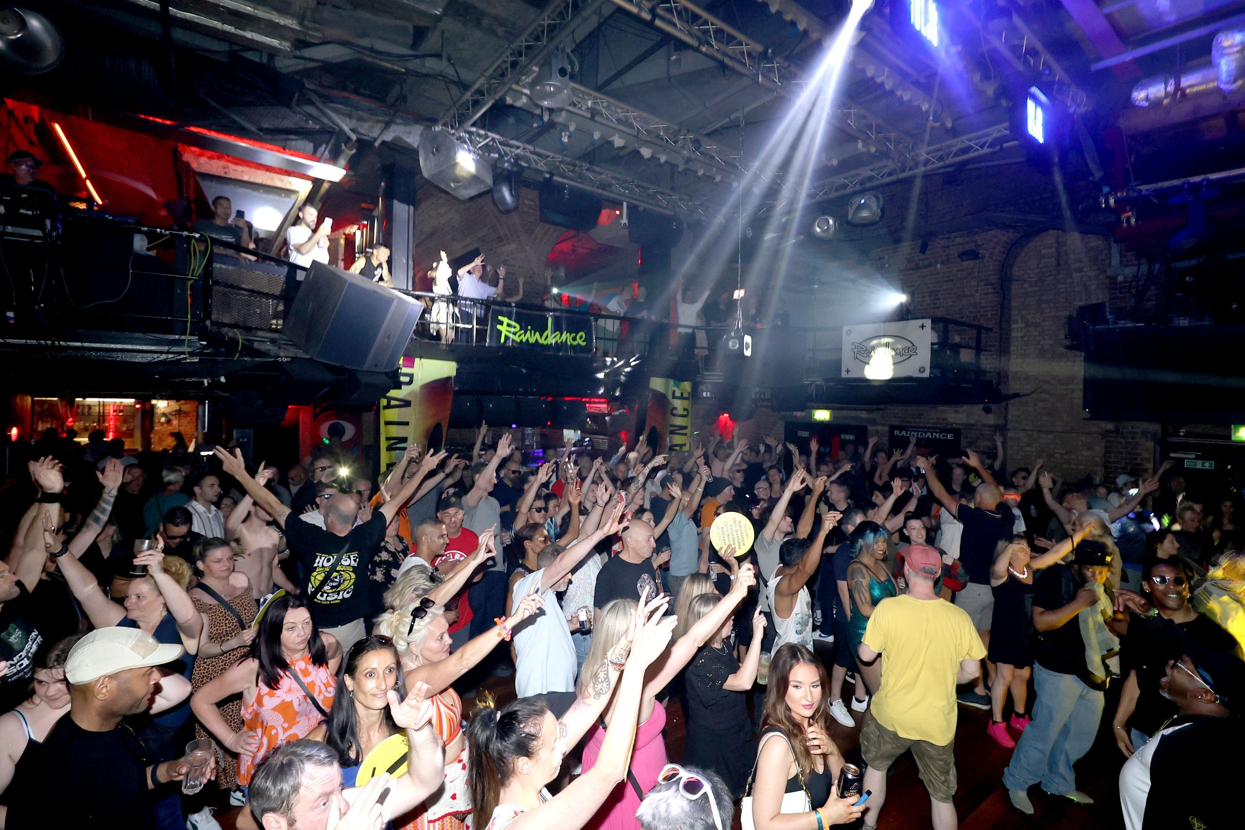 Crowd of people at a dance club with raised hands, dancing and socializing under colorful lights and a high ceiling. There is a DJ booth on an upper level with people, and large speakers and lighting equipment surround the scene.