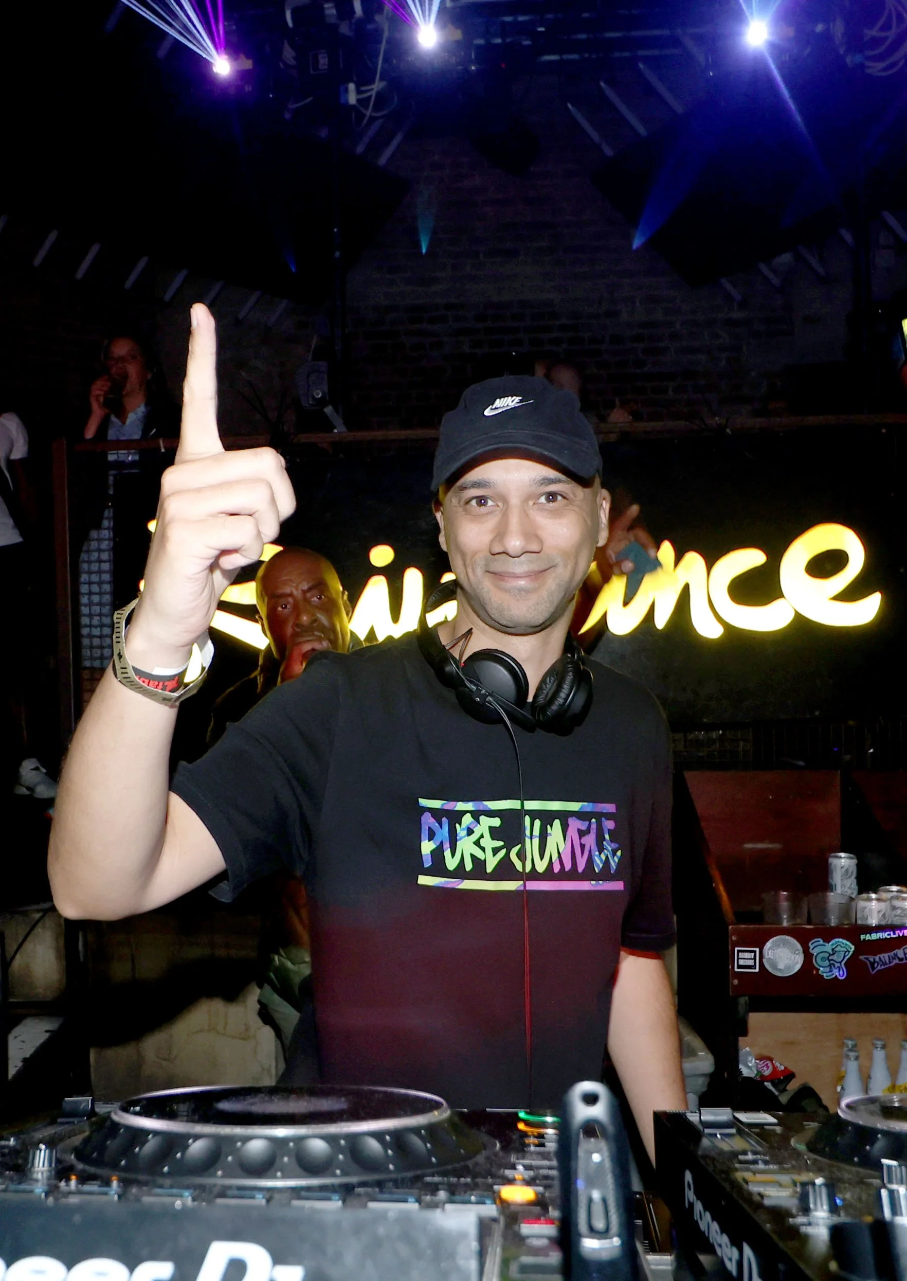A DJ at a club or event, smiling and raising one finger, wearing a black Nike cap and a "Pure Jungle" T-shirt with headphones around his neck, with a neon "Raindance" sign in the background.