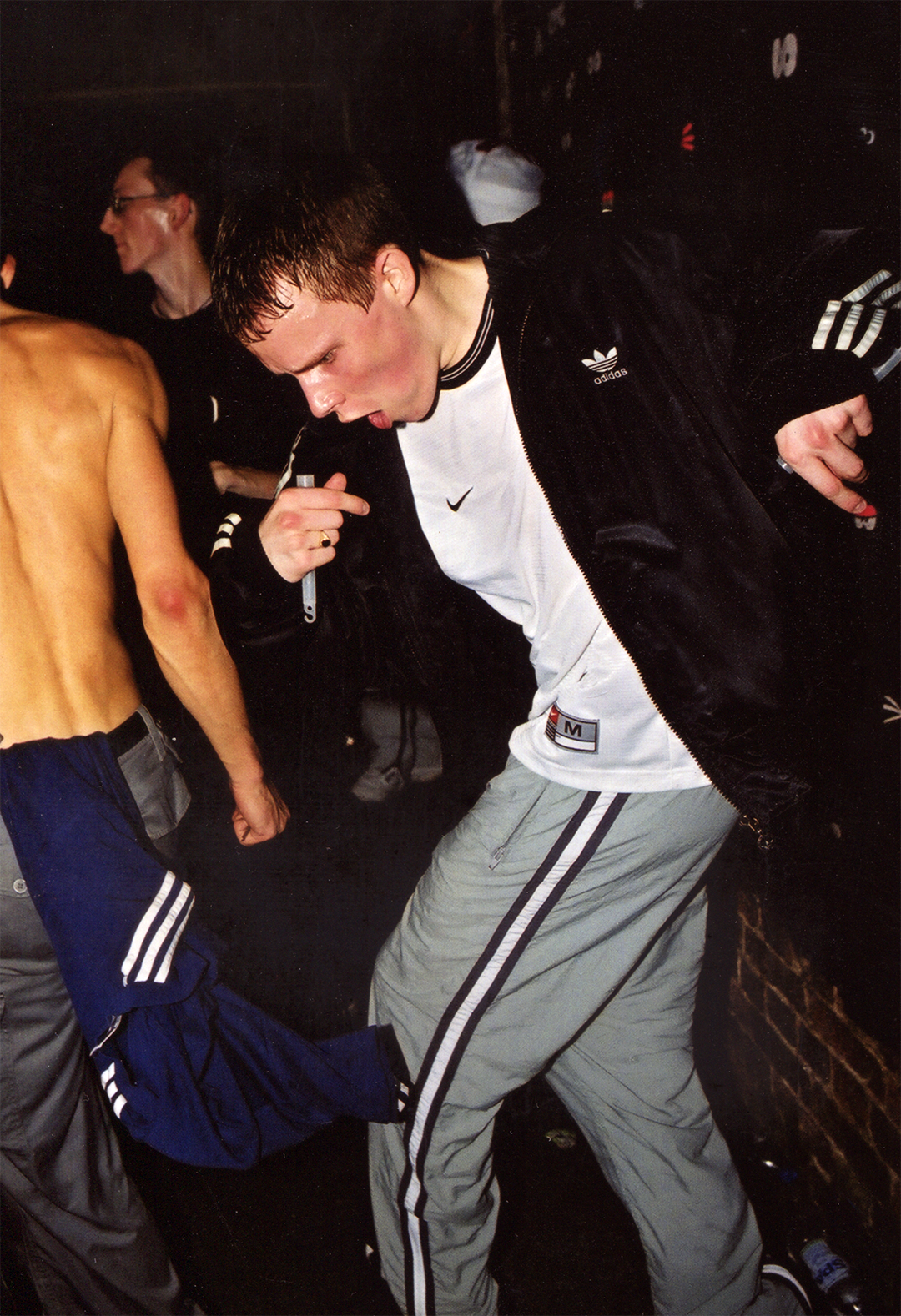 A young man in a white Nike shirt and gray Adidas track pants, with a black Adidas jacket, is dancing or moving to music in a dark room. He has short brown hair and is making a fist with his right hand. There are other people in the background, including a shirtless man and a woman with glasses. The setting appears to be a lively party or club.