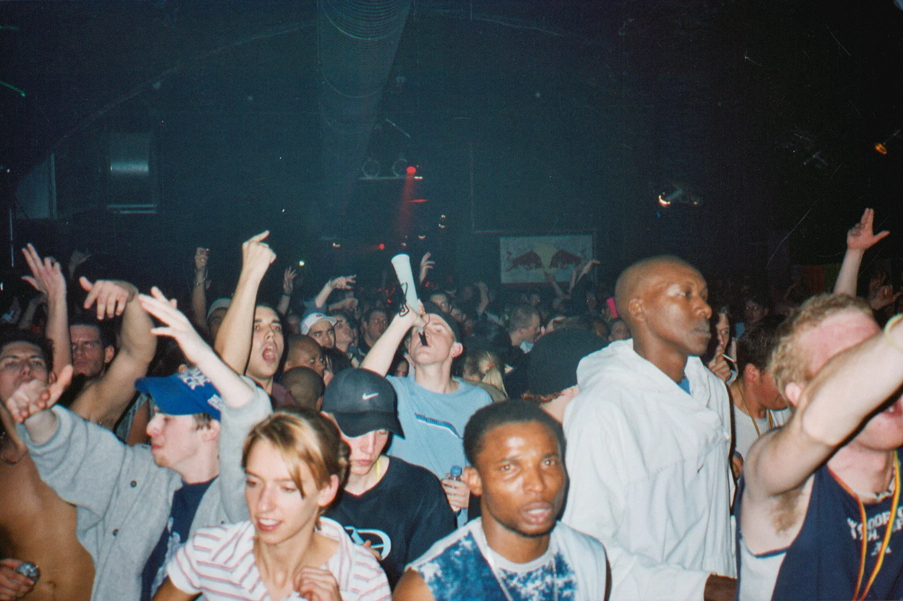 Crowd of people dancing and enjoying music at a concert or nightclub with dark lighting and some audience members raising their hands.