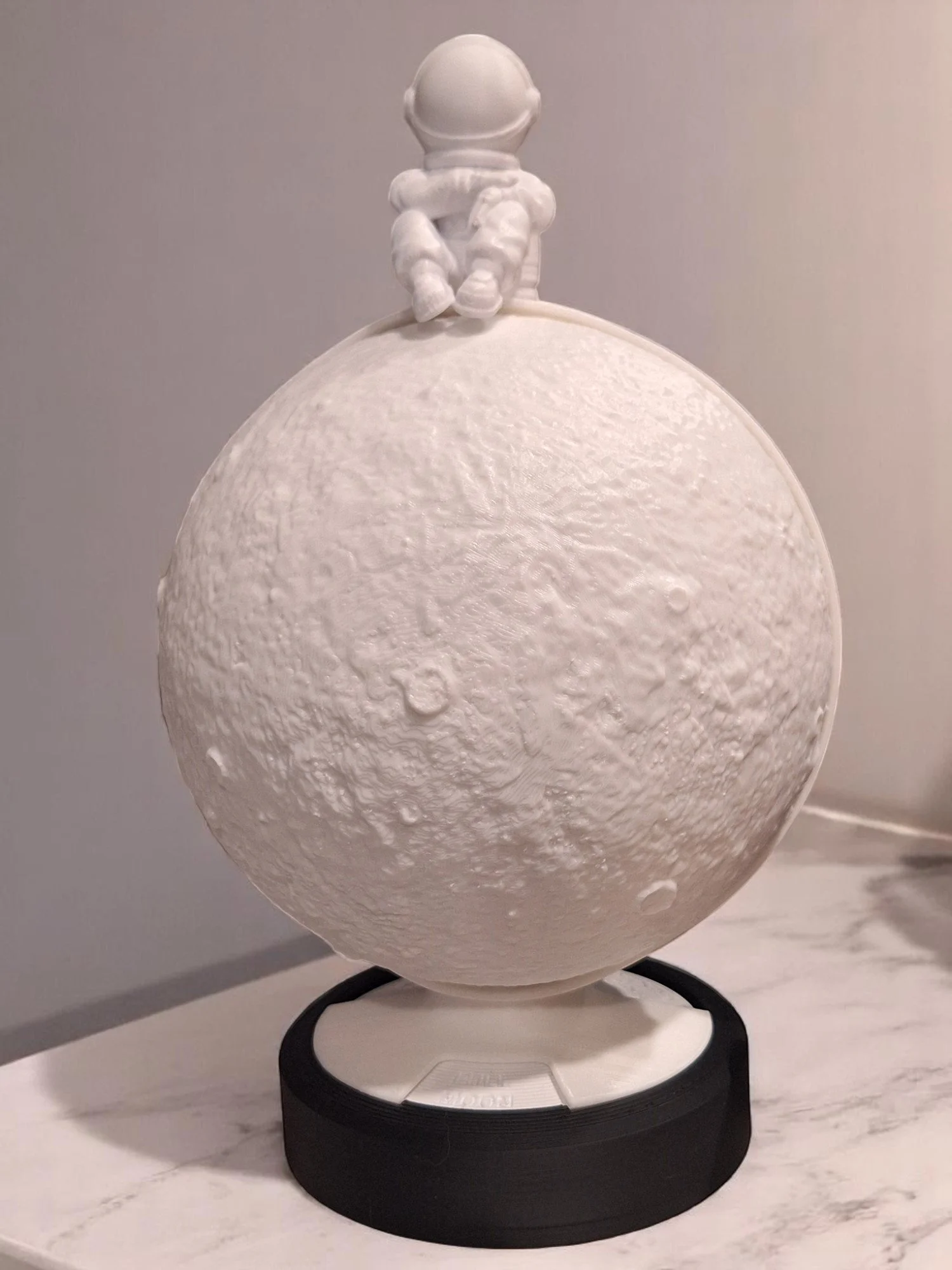 A white 3D-printed model of the Moon with an astronaut figure sitting on top, placed on a black and white circular platform, on a white marbled surface.