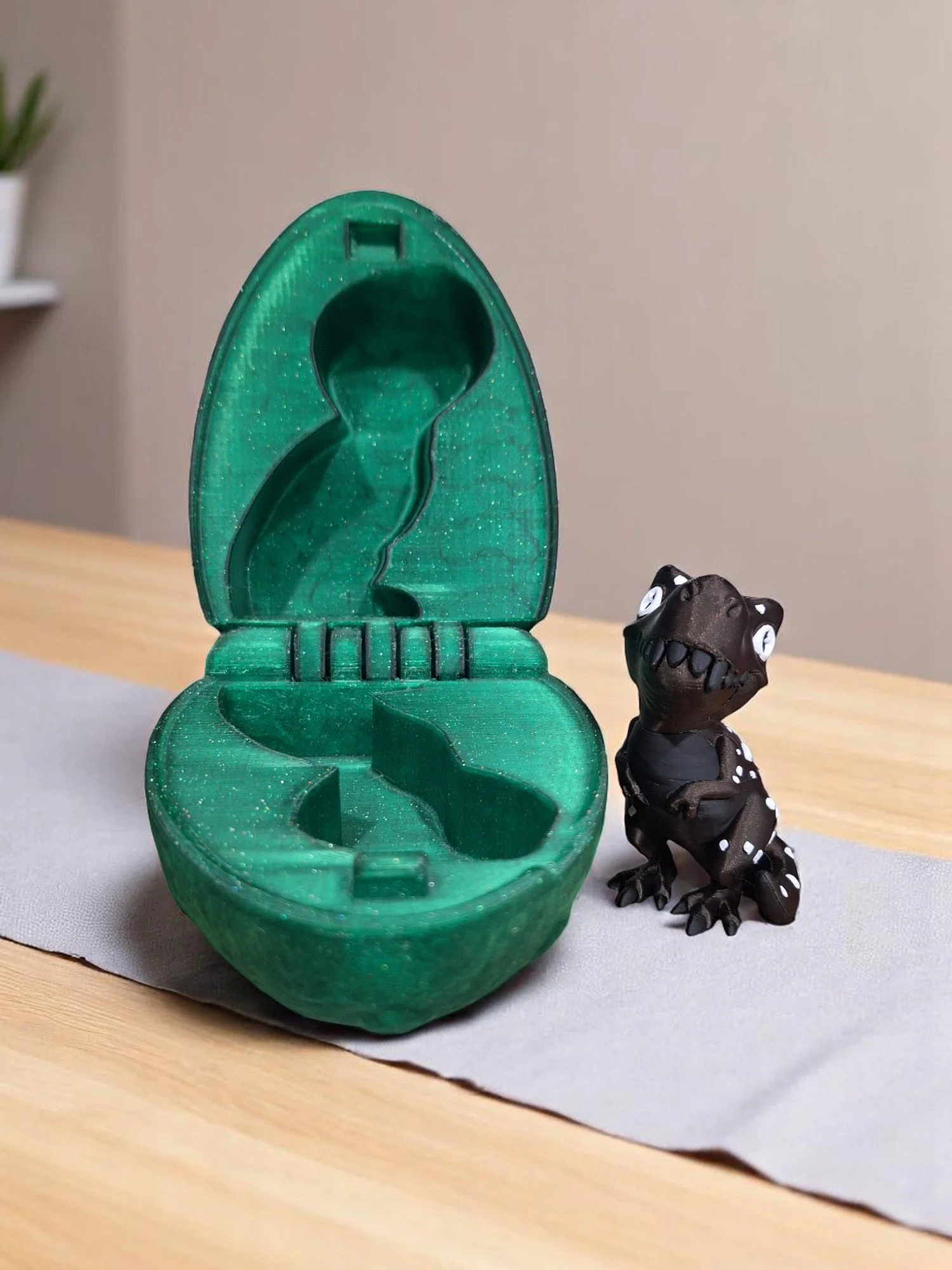 A green, dinosaur-shaped container with a removable lid next to a small black toy dinosaur with white spots on its back, sitting on a light gray mat on a wooden table.
