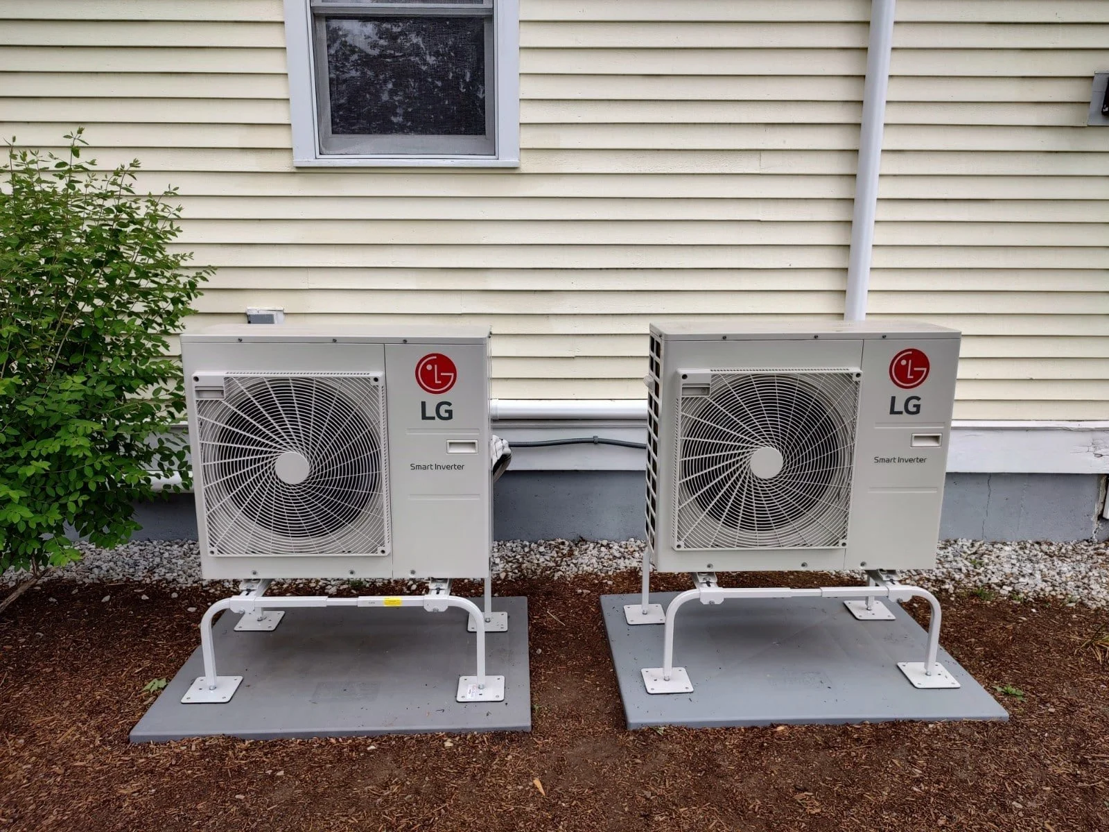 Two outdoor LG smart inverter air conditioning units installed on concrete pads outside a house with beige siding and a window.