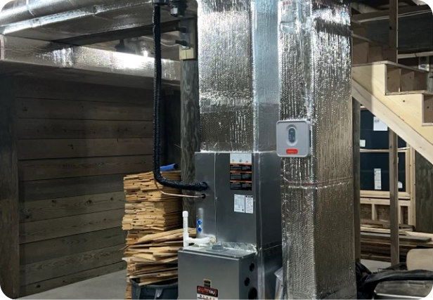 An HVAC furnace installed in a basement or utility room with wooden walls, with a stack of wood planks nearby and stairs leading up on the right side.