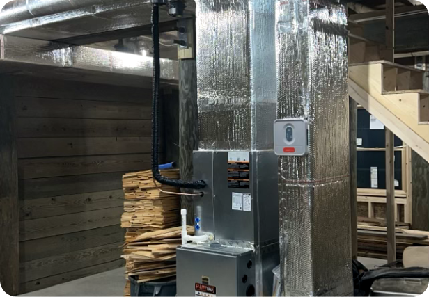 A heating, ventilation, and air conditioning (HVAC) air handler with silver insulation, situated in an unfinished basement with wooden stairs and stacked wooden planks in the background.