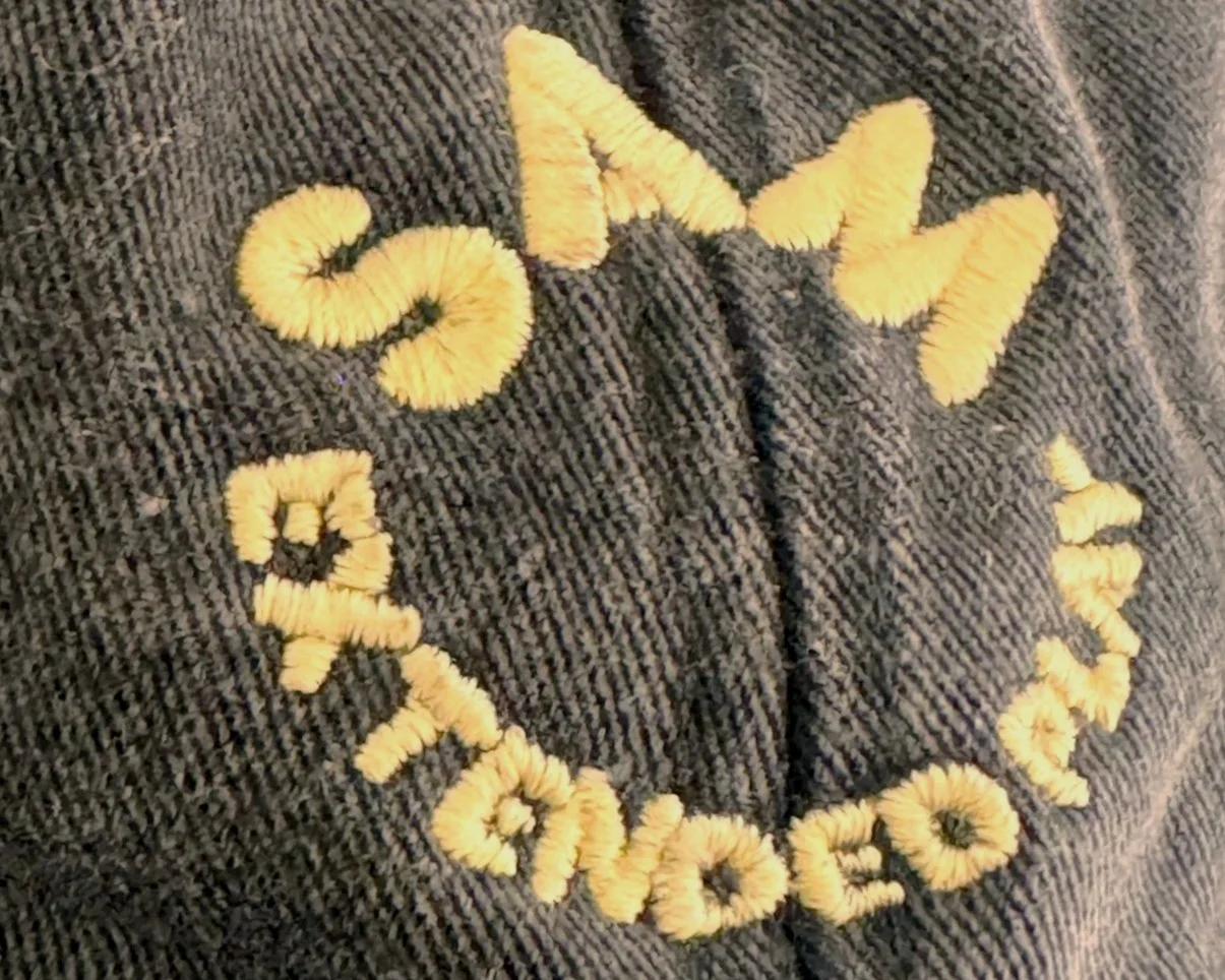 Close-up of dark fabric with embroidered yellow text that reads 'SAM' 'EXTENDED PLAY'.