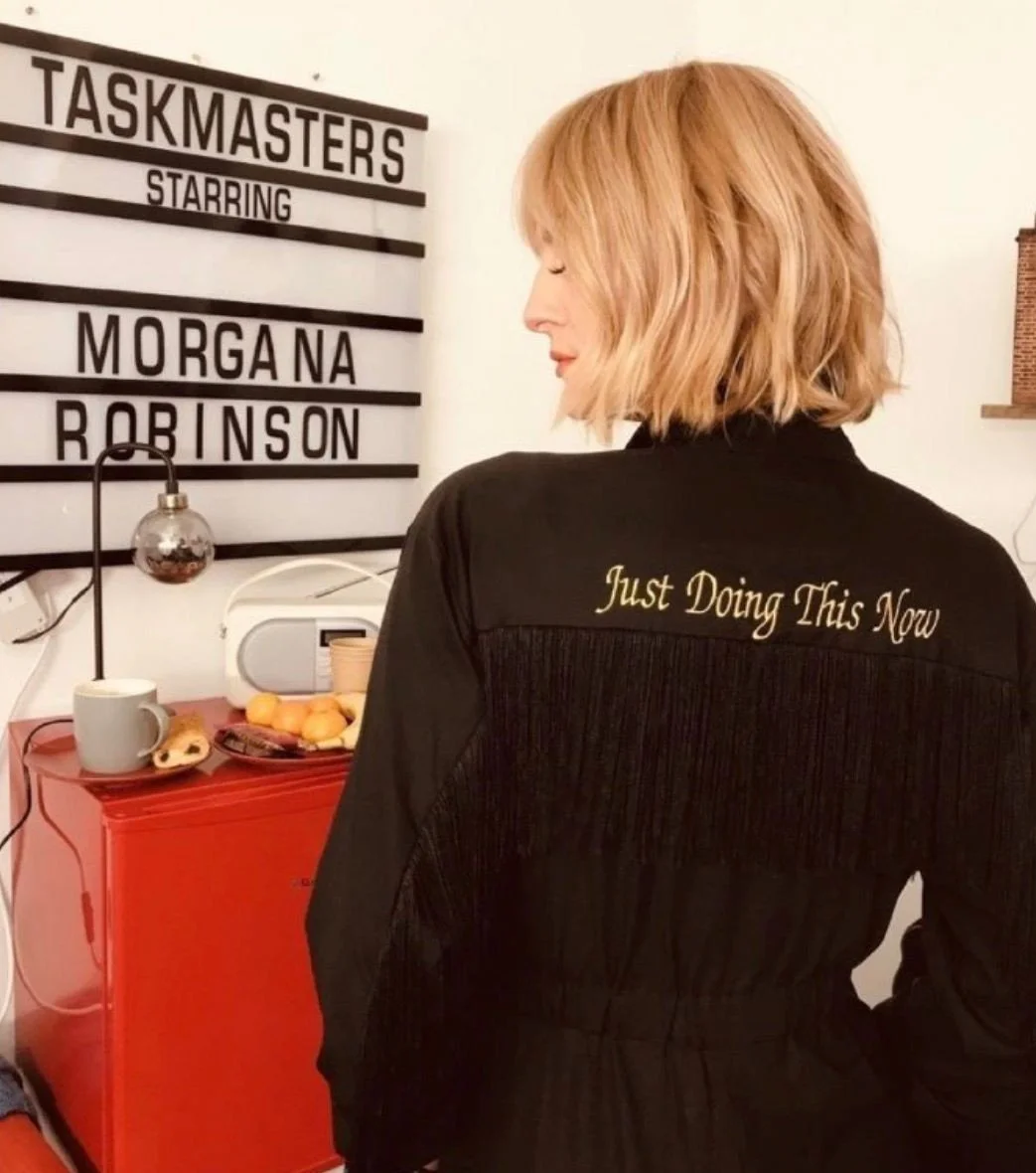 A woman with blonde hair standing in front of a sign that reads "TASKMASTERS STARRING MORGANA ROBINSON". She is wearing a black jacket with the words "Just Doing This Now" embroidered on the back. 