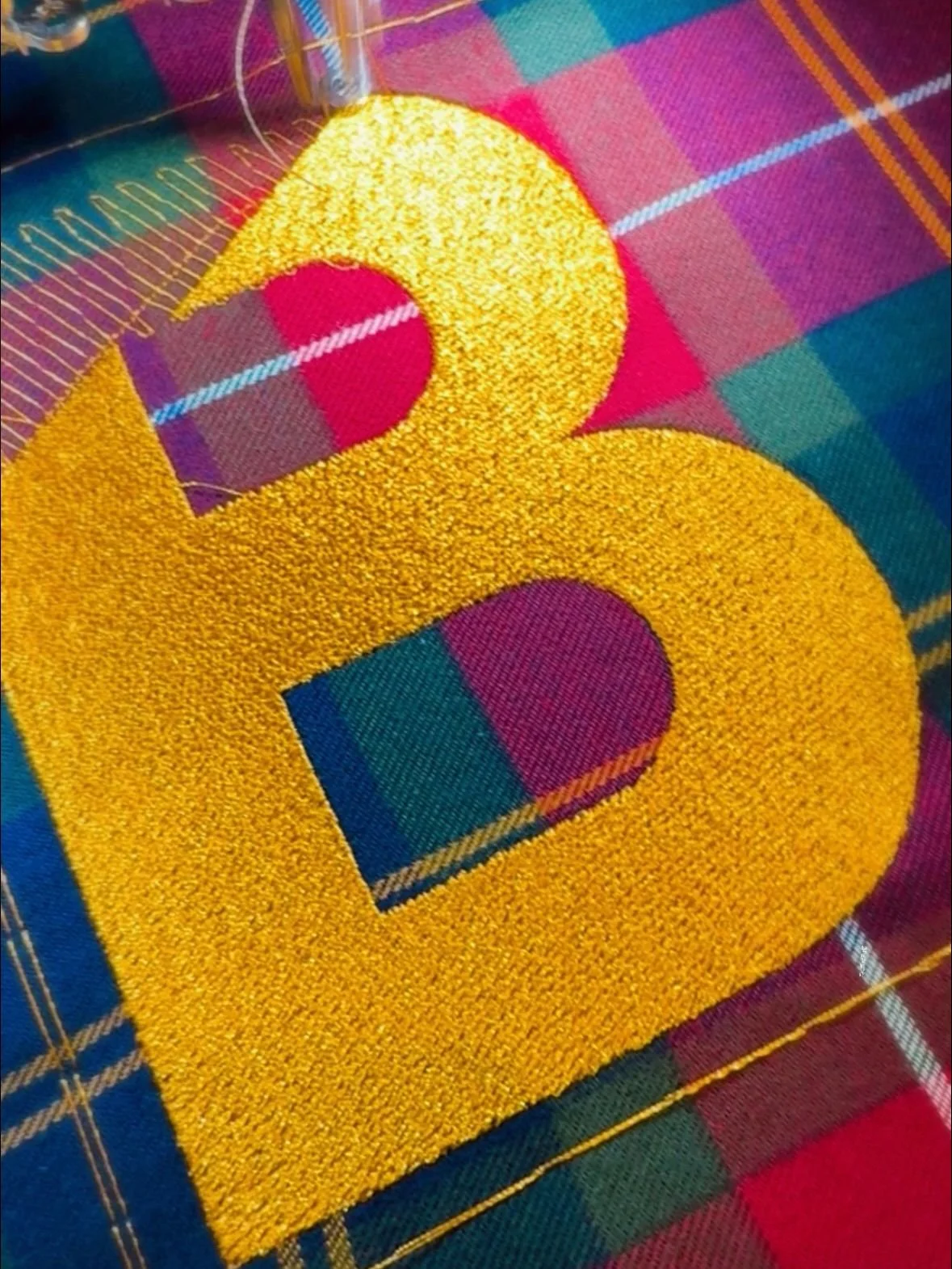 Close-up of a stitched letter 'B' in yellow thread on colorful fabric with a plaid pattern.
