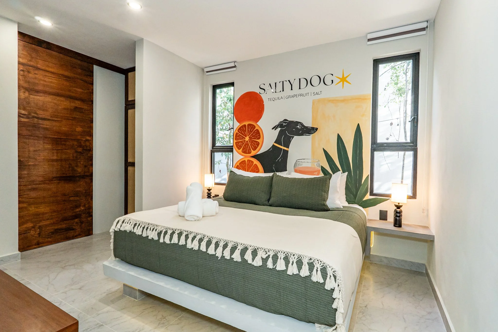 A bedroom with a white bed, green bedspread, and white tassel blanket, two rolled towels on the bed, wood paneling on the left wall, a large mural of a saluki dog with citrus slices, the word SALTYDOG, and ingredients tequila, grapefruit, and salt, two windows, and two bedside lamps.