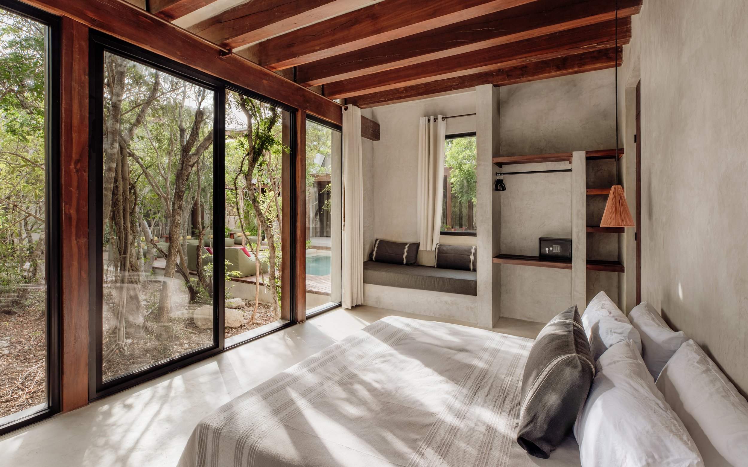 A bedroom with large sliding glass doors opened to a garden with trees. The room has wooden beams, a small window with white curtains, a built-in window seat with cushions, and a bed with white and black bedding. There's also a small nightstand with a lamp and a safe on the wall.